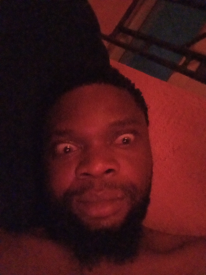 The image is a close-up, chest-up shot of a person, likely male, with dark skin, dark hair, and a dark beard. The person is looking directly at the camera with wide, open eyes, showing a significant amount of white around the irises. Their mouth is slightly open or in a relaxed, somewhat pursed position. The overall lighting is dominated by a strong, reddish-orange hue, casting a warm, almost intense glow on the subject's face and the surrounding environment.
The person's facial expression appears to convey surprise, shock, or intense focus due to the wide-eyed gaze. Their dark, short, possibly curly hair is visible around the forehead and temples, and a full beard covers their chin and jawline. The upper part of their chest is visible, suggesting they might be shirtless or wearing very dark clothing that blends with the shadows.
The background is largely out of focus due to the close proximity of the subject to the camera. On the right side, a reddish wall or surface is visible, matching the dominant lighting. In the upper right corner, some dark, geometric shapes with faint lighter outlines can be discerned, possibly indicating metal bunk bed frames or shelves, partially illuminated by the same reddish light. The left side and upper left portion of the image are considerably darker, almost black, providing a strong contrast to the illuminated right side and the subject's face. The image has a slightly grainy quality. No text is visible in the image.