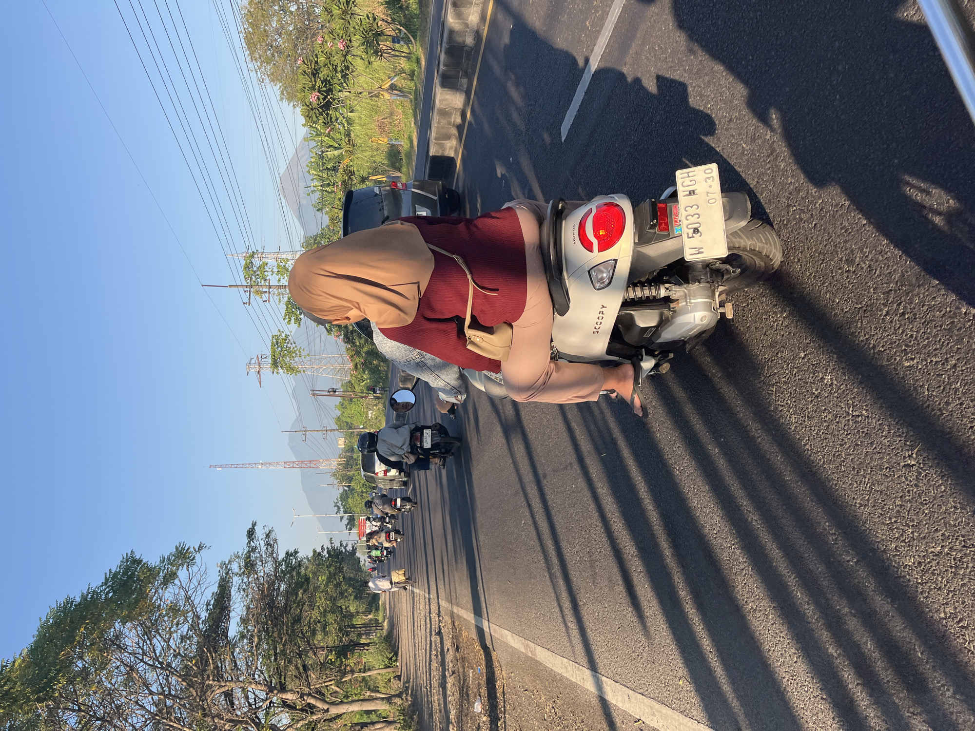 In the city of Siring, Indonesia, a person wearing a hijab and a maroon sweater is riding on the back of a white Honda Scoopy motorcycle on a paved road. The motorcycle has a license plate that reads "W 5033 NGH" and "07-30". The rider is wearing tan pants and black flip-flops. The scene is set during the day, with a clear blue sky and sunlight casting long shadows from the motorcycle and other vehicles on the road. Other motorcycles and a black SUV are also visible on the road, indicating traffic. Trees line the side of the road, and in the background, mountains and electrical towers can be seen, suggesting a semi-urban or rural setting. The overall impression is one of everyday life and transportation in Indonesia.