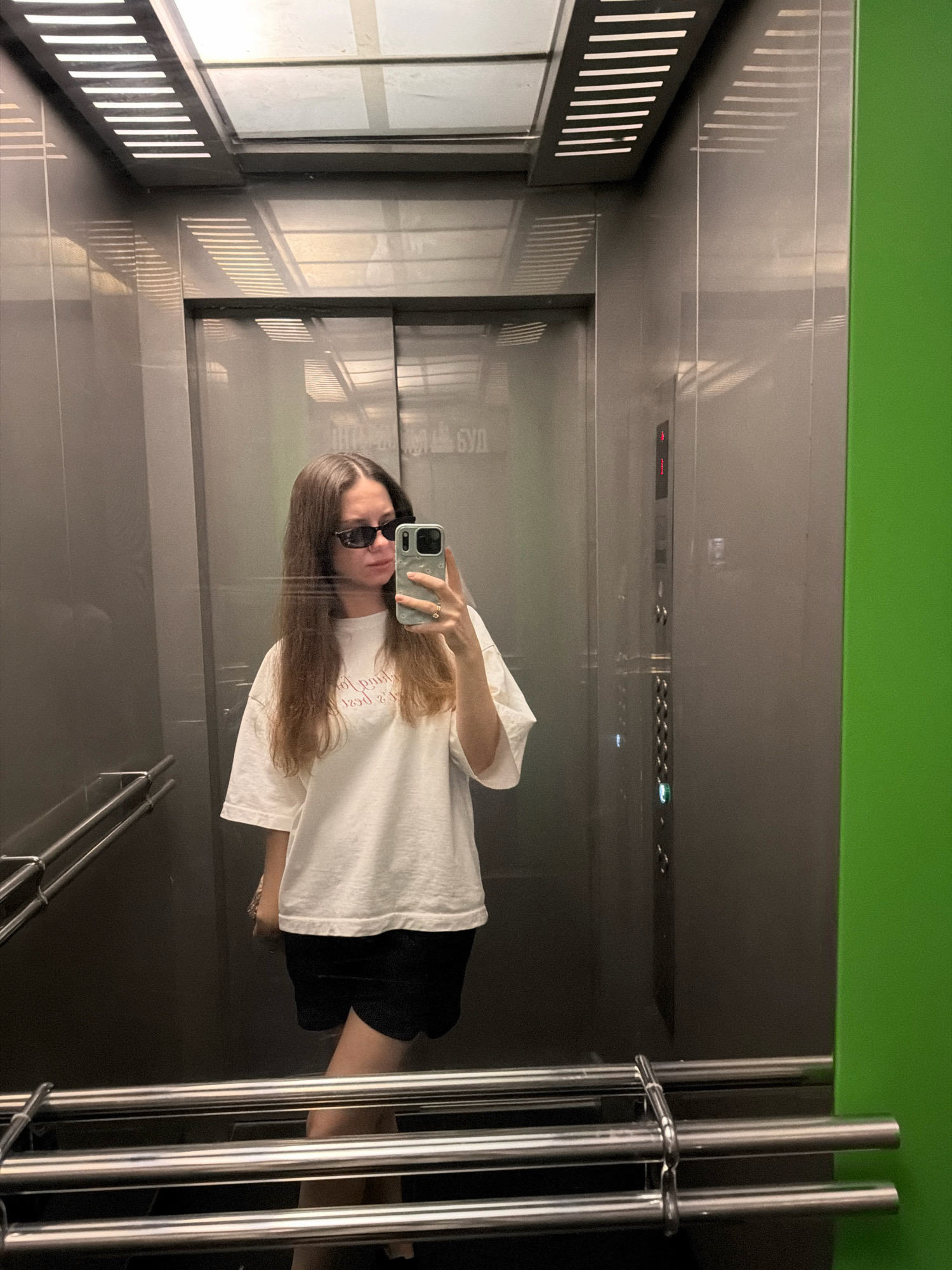 A young woman with long brown hair and sunglasses takes a selfie in an elevator. She is wearing a white t-shirt and black shorts. The elevator has silver walls and a green panel on the right side. There are handrails on the left side of the elevator. The ceiling has a light fixture with horizontal white slats. A phone is visible in her hand as she takes the picture. The image appears to be taken indoors, possibly in a residential building or a public space within the city of Kyiv, Ukraine. The lighting is artificial, casting reflections on the metallic surfaces of the elevator.
