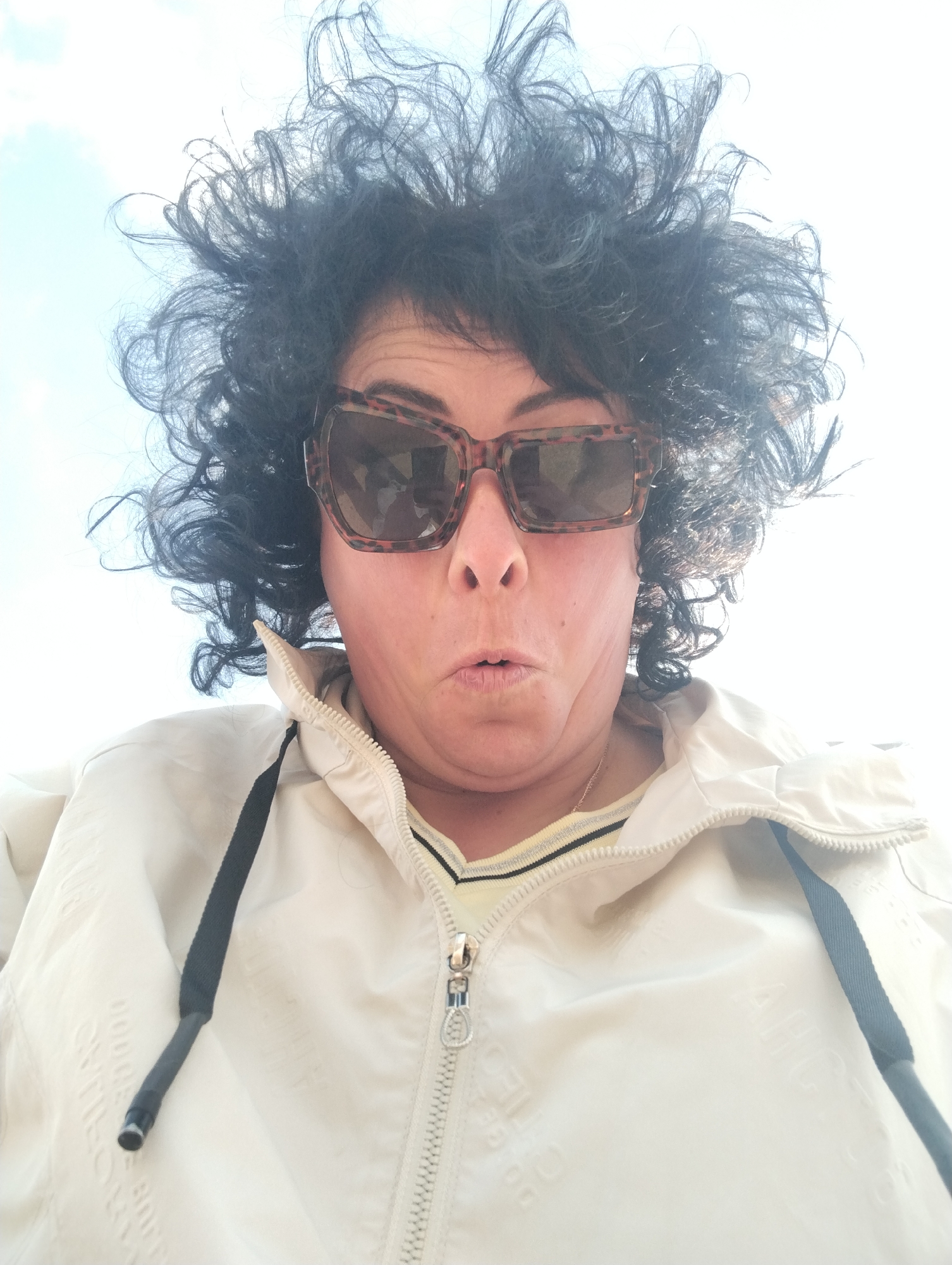 A woman with dark, frizzy hair, wearing leopard-print sunglasses and a light-colored windbreaker, is looking down at the camera with a surprised expression. The background is bright, suggesting outdoor lighting and potentially a sky above. No information suggests this image was taken in the city of Grishanki, Russia.