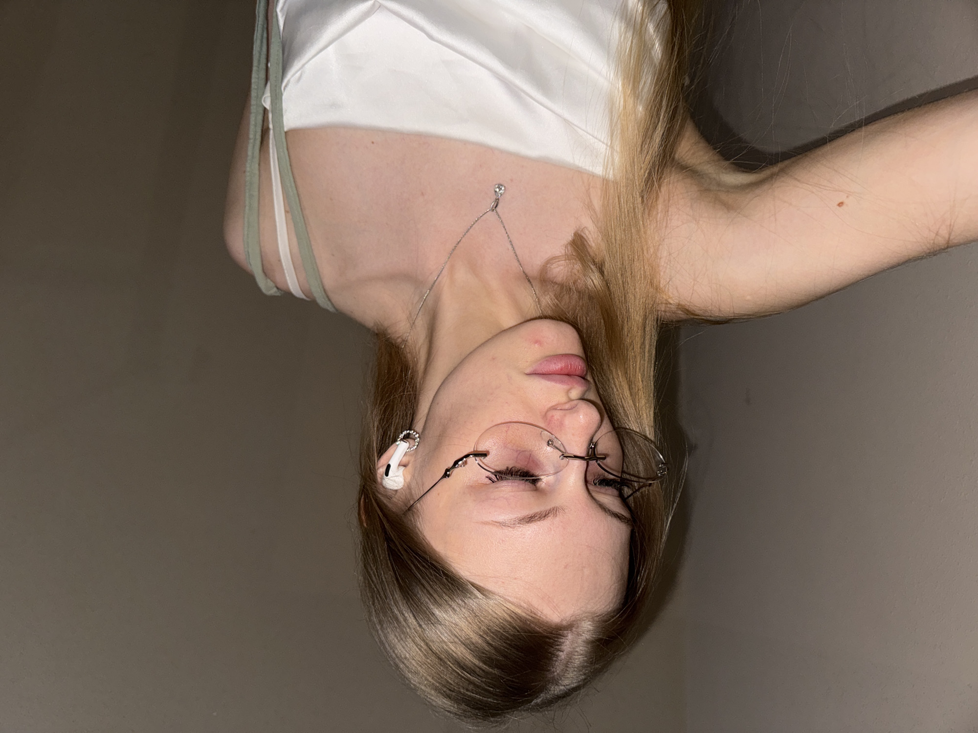 The image is a close-up, rotated shot of a young woman. She has long, blonde hair and is wearing a white, silky top with thin straps. She is also wearing glasses with thin, rimless frames, and white earbuds in her ears. A delicate silver necklace with a small pendant is visible around her neck. Her eyes are closed, and her lips are slightly parted, suggesting a relaxed or pensive mood. The background is a plain, neutral wall, indicating an indoor setting. The lighting is dim, creating shadows and a soft focus on the subject. There is no visible text in the image. The setting is likely domestic, given the casual attire and indoor environment. The time of day is not discernible due to the artificial lighting and lack of external cues. The overall mood is intimate and serene.