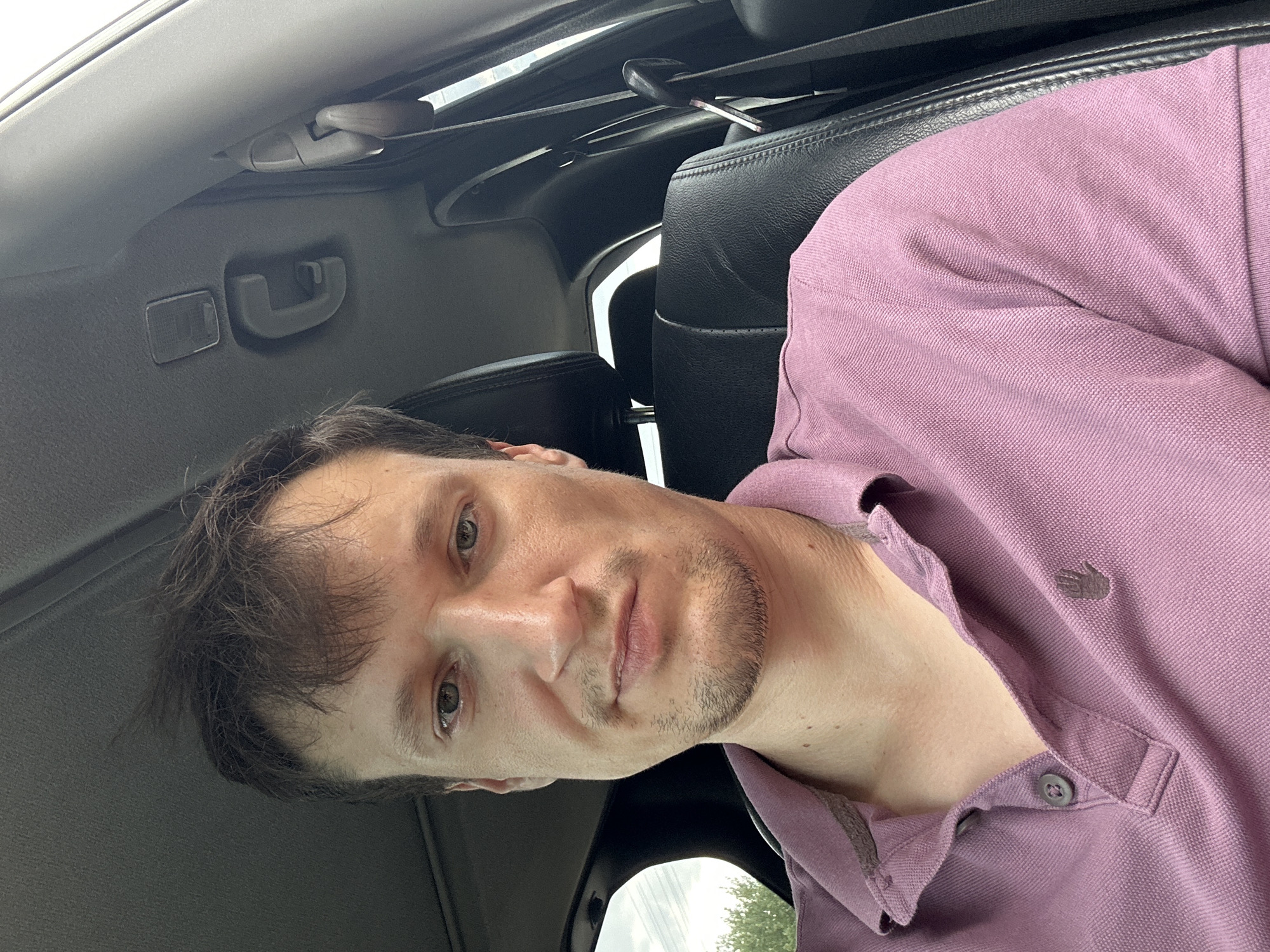 A man is sitting in the driver's seat of a car, looking at the camera. The man has short brown hair and is wearing a pink polo shirt. The interior of the car is visible, including the black leather seats and the car's ceiling. The window on the left side of the car shows a blurry background of trees and sky, suggesting the car is outdoors. The overall lighting indicates it is daytime. The image does not provide any specific location information within the city of Makiv, Ukraine.