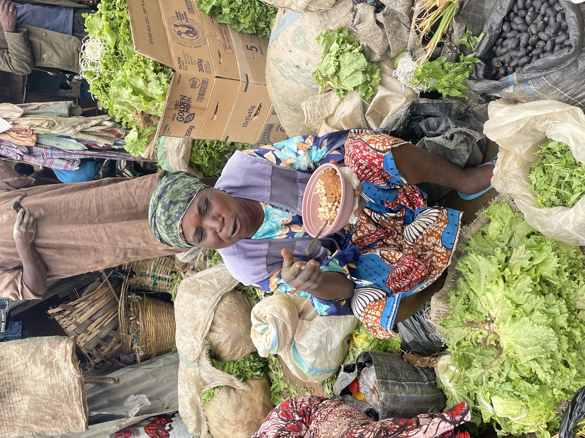 This media file shows a top-down view of a market scene, likely in Gura Top, Nigeria. The primary subject is a person in the center, wearing a green headscarf and a colorful patterned dress. They are holding a small bowl containing food and a spoon, appearing to be eating. Around this person, the scene is filled with various agricultural goods. Prominently featured are large bunches of lettuce, some of which are in cardboard boxes with visible text, including brand names like "GOLDEN MORN" and weight indications such as "300g". There are also large sacks containing dark, olive-like produce, and bags filled with green leafy vegetables. Woven baskets are also visible, suggesting the retail nature of the environment. The lighting and the type of produce indicate it is daytime, and the bustling market atmosphere is palpable. The person's expression is relaxed, possibly indicating a moment of rest or a meal break during a busy market day. The overall impression is one of a vibrant local food market.