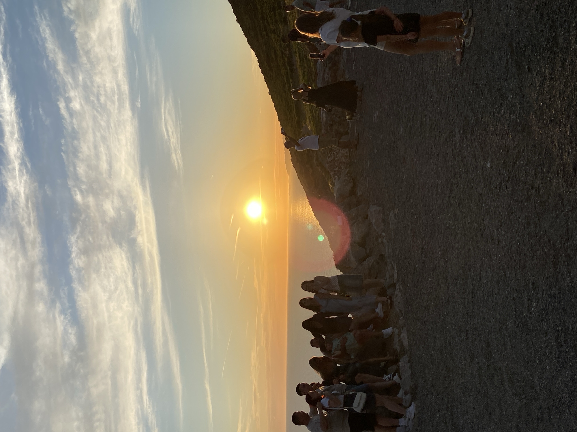 The image depicts a group of people gathered on a rocky terrain overlooking the sea at sunset. The sun is low on the horizon, casting a warm golden light across the scene and reflecting on the water. Some individuals are standing, while others are seated. Several people are holding up their phones, seemingly capturing the moment. A man in a white shirt and khaki shorts is standing and looking towards the sun. In the foreground, two young girls are visible, one with her arm around the other. The environment is characterized by rugged, rocky ground and a sloping hillside covered in vegetation in the background. The sky is a blend of orange, yellow, and blue, with wispy clouds. The location is identified as Kovačko Polje, Montenegro.