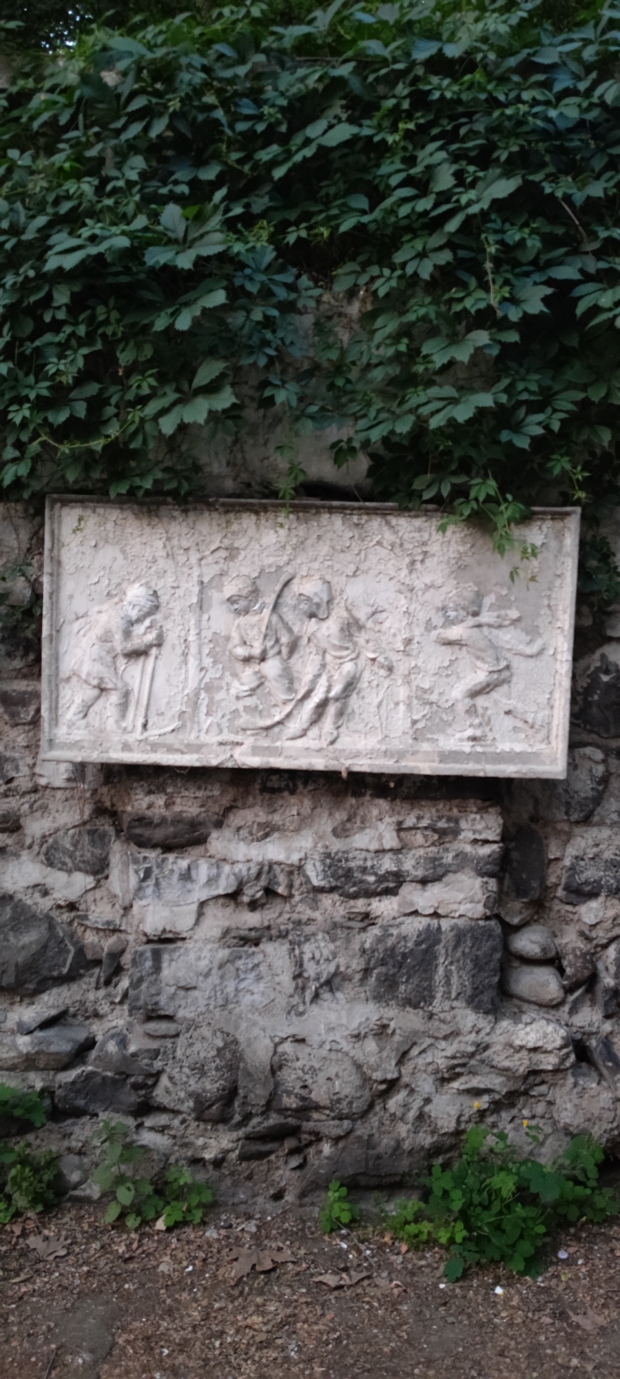 The image displays a bas-relief sculpture mounted on a rough stone wall, overgrown with green ivy. The sculpture, a rectangular panel, depicts three figures in what appears to be a narrative scene. On the left, a figure stands with a tool, possibly a shovel or pickaxe, held upright. In the center, two figures are huddled together, one bending slightly. To the right, a third figure is depicted in motion, possibly running or dancing, with arms outstretched. The sculpture is weathered and shows signs of age, with chipped and peeling surfaces. The stone wall beneath is constructed of irregularly shaped stones of varying sizes and colors, some of which are partially covered by dirt and small plants. The ground in front of the wall is composed of gravel, dirt, and scattered dry leaves. The overall setting suggests an outdoor environment, possibly a garden or an older section of a city. This sculpture is located in the Matenadaran museum complex in Yerevan, Armenia, specifically in the courtyard area.
