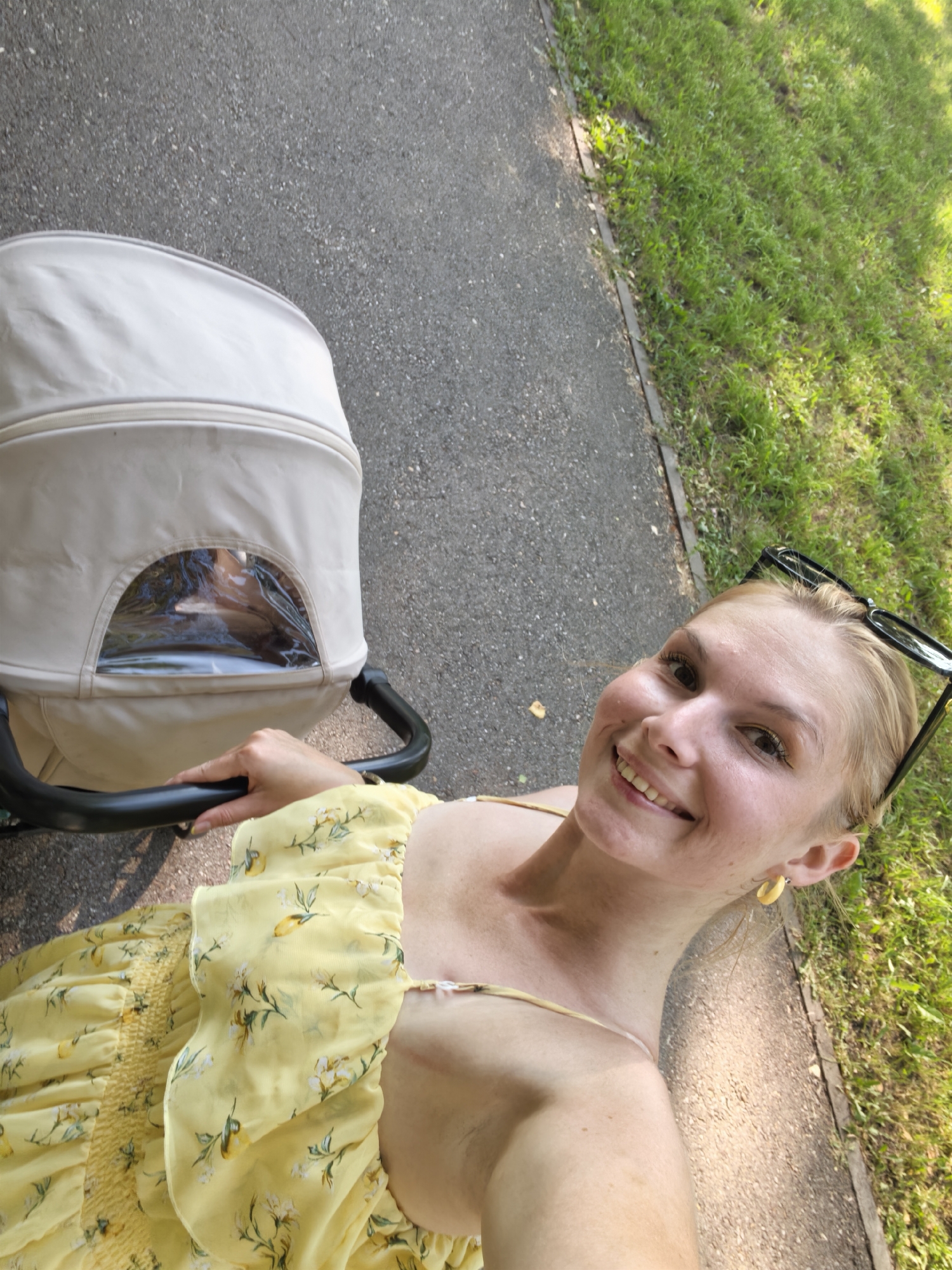 In this tilted, outdoor, medium shot, a blonde woman with fair skin is captured from the chest up, holding a stroller. She is wearing a yellow floral dress and yellow hoop earrings, with dark-rimmed sunglasses perched on her head. Her eyes are brown, and she is smiling broadly, revealing her teeth. The stroller, positioned to her left, is light-colored and appears to be made of fabric, with a transparent window on its side. The background consists of a grey asphalt path and a grassy area with green grass, indicating an outdoor setting, possibly a park or a residential area. The lighting suggests it is daytime, with sunlight creating shadows on the path. The image does not contain enough information to ascertain the specific location within the city of Sterlitamak, Russia.