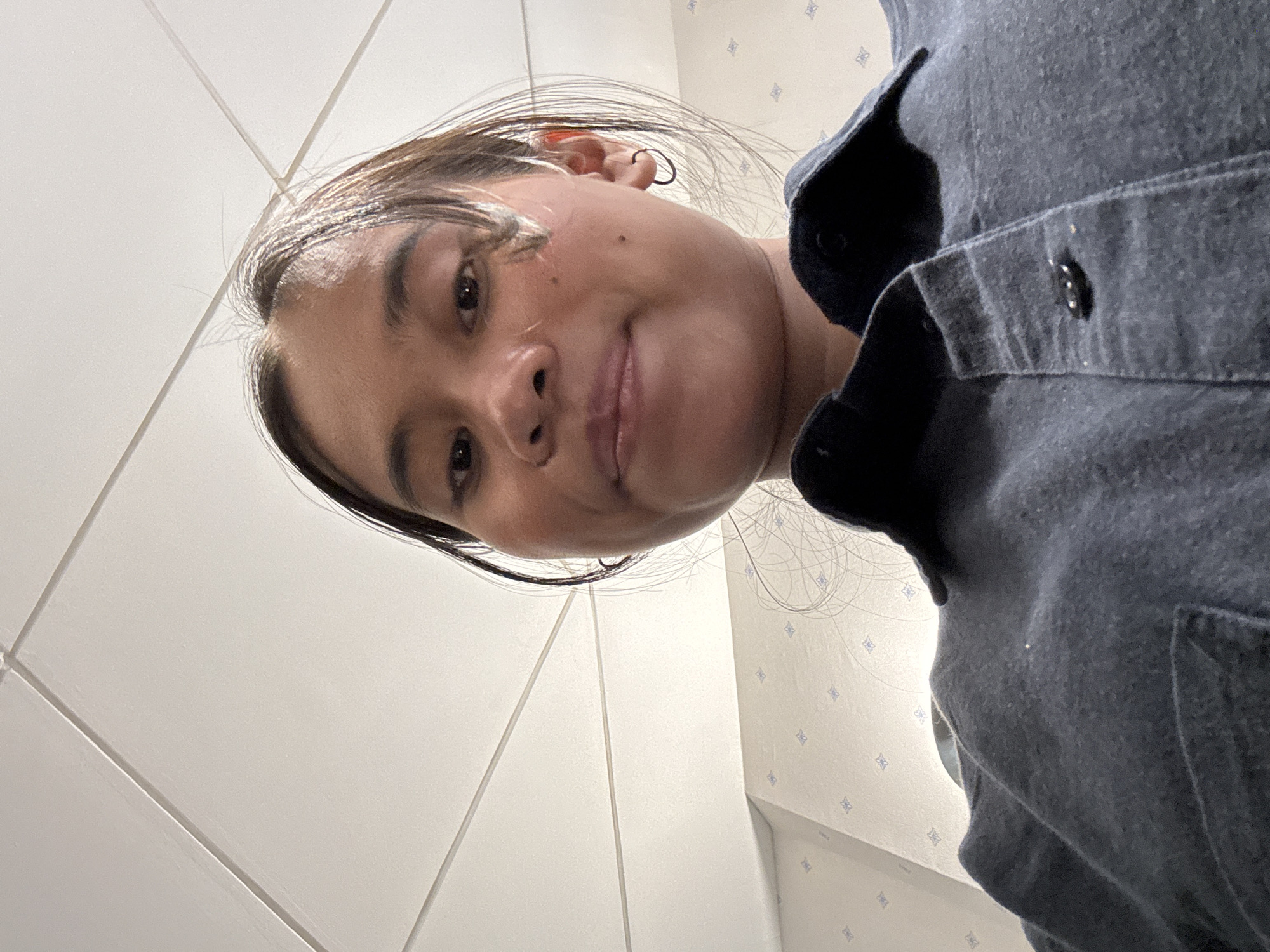 An overhead, slightly distorted, close-up, chest-up selfie of a young woman with medium-dark skin and long, dark brown hair. She is wearing a dark blue collared shirt and a hoop earring in her left ear. She has a small mole on her left cheek. The background consists of a white tiled ceiling and a wall with a light beige wallpaper featuring a subtle blue diamond pattern. The photo appears to have been taken indoors, possibly in a residential or office setting. There is no discernible information in the image to indicate a specific location within South Jakarta, Indonesia.