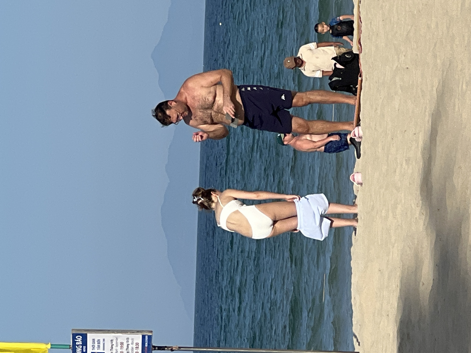 The image shows a beach scene in Đà Nẵng, Vietnam, with several people visible. In the foreground, a man in dark blue swim trunks is standing and appears to be engaged in some activity, possibly stretching or exercising. To his right, a woman in a white bikini is also standing, with a light blue garment draped over her back. The background features the ocean with gentle waves and a hazy sky, suggesting a clear day. Further in the background, other individuals can be seen on the beach, some standing and others potentially sitting or crouching. One man is wearing a light-colored shirt and a cap, while another person in the distance is wearing a dark top. The lighting suggests it could be daytime, possibly late morning or afternoon. A sign with Vietnamese text is partially visible in the lower left corner, which is common for beach information or regulations. The overall mood appears relaxed and casual, typical of a beach environment. The presence of mountains in the far distance further confirms the coastal setting.