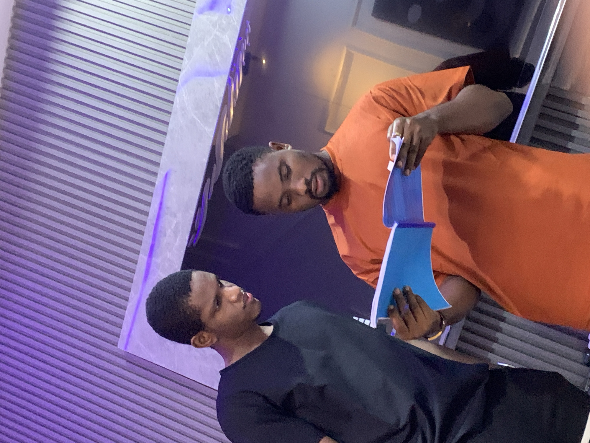 In a location identified as Igboefon, Nigeria, two adult males are observed standing indoors, focused on a shared object. The male on the right, wearing an orange short-sleeved t-shirt and displaying a beard, holds an open blue notebook or book with visible white pages, appearing to read or examine its contents. A gold-colored ring is present on his left ring finger. The male on the left, wearing a black short-sleeved t-shirt, is positioned slightly behind and to the left of the first individual, observing the same open notebook. The background consists of a vertically corrugated or ridged wall section, illuminated by purple ambient lighting, and a smooth, possibly reflective, horizontal surface also accented with purple light. The overall environment suggests a modern indoor setting.