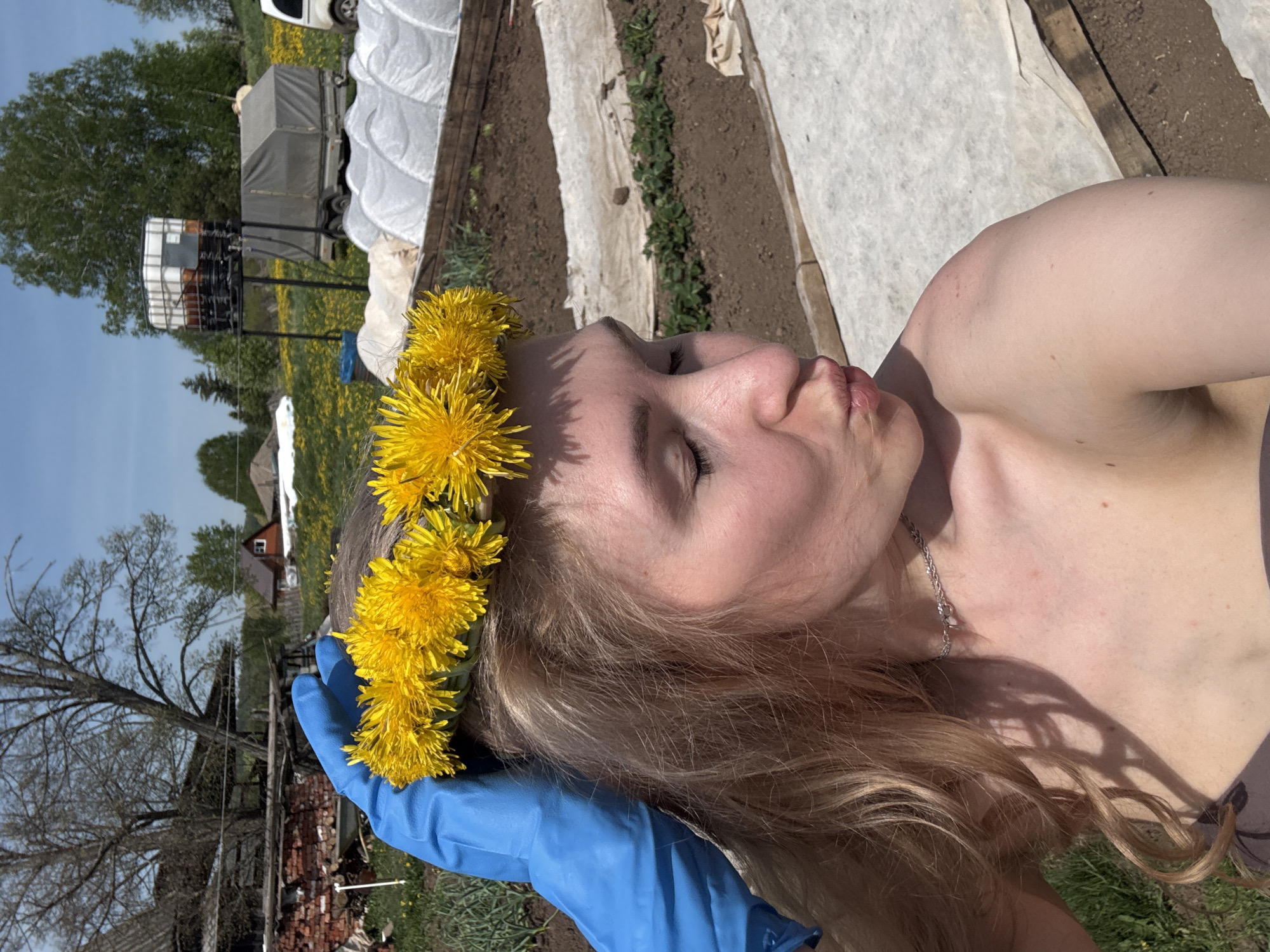A woman wearing a dandelion crown and blue surgical glove poses outdoors in Nizhnyaya Chermoda, Russia. The backdrop features agricultural elements such as rows of cultivated beds covered with white fabric, potentially in a garden or farm setting. Additional details include a trailer with a cover, bare trees and a blue sky suggesting a spring or early summer season. A partially ruined brick structure and what appears to be a rural dwelling are also visible.