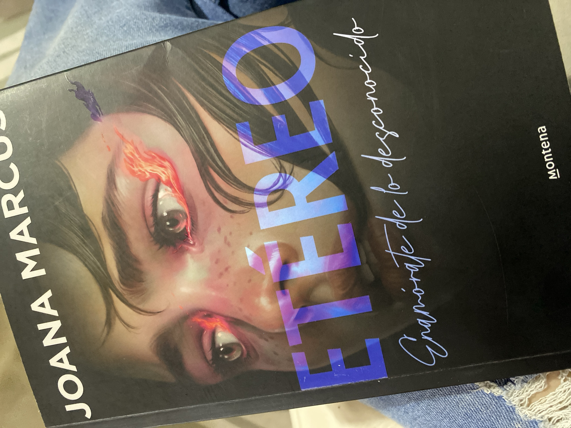 The image is a close-up of a book cover, featuring a stylized illustration of a person's face, primarily focusing on the eyes. The title, "ETÉREO," is prominently displayed in large, iridescent blue letters across the lower half of the face. Above it, in white block letters, is "JOANA MARCUS." Below "ETÉREO," a script text reads "Enamórate de lo desconocido." In the bottom right corner, the word "MONTENA" is written in white, underlined. The background is black, and the book is resting on what appears to be blue denim, possibly jeans. There is no visible indication of the city of Macho Muerto, Venezuela, within the image.