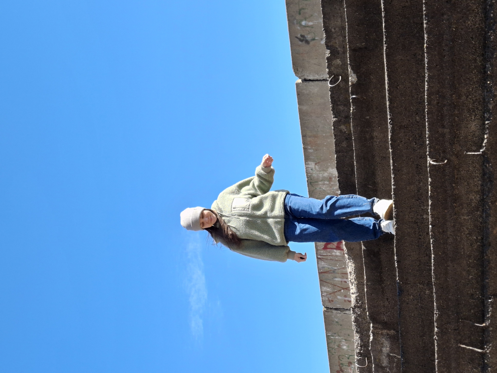 The image features an upward-angled shot of a young person standing on a weathered concrete structure against a bright blue sky with a faint cloud. The subject is positioned on one of the ledges of a multi-tiered concrete structure, looking slightly downwards with a soft smile. They wear a light green fleece jacket, blue jeans, white sneakers, and a light grey beanie, with long dark hair falling from beneath the beanie. One arm is slightly raised, possibly for balance or gesture.
The concrete structure consists of several distinct horizontal layers or ledges, with metal loops embedded in places. Faint graffiti marks are visible on some sections of the concrete. The low camera angle emphasizes the structure's height and the subject's elevated stance. The bright, sunny conditions indicate daytime. This scene is located in Chaikovsky, Russia. No readable text is visible.