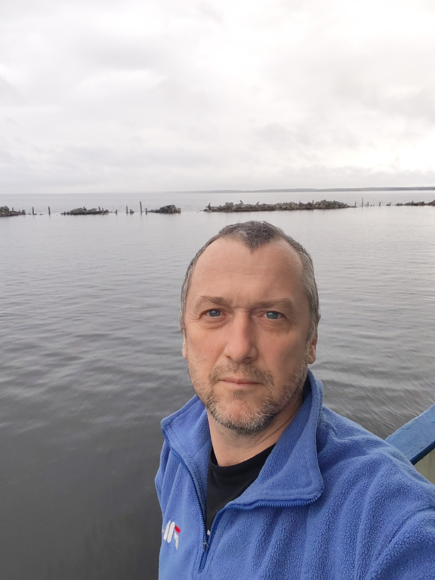 A middle-aged man is captured in a selfie, looking directly at the camera with a neutral and calm expression. He has blue eyes, a short beard, and greyish hair. He is wearing a blue fleece jacket with a stylized "NR" or similar logo in white and red on the left chest, over a black t-shirt. A blue railing or structure is partially visible on the right side of the frame, suggesting he is on a boat, pier, or bridge.
The setting is outdoors, at what appears to be a large body of water, possibly a lake or bay, under a heavily overcast, light grey sky. The water is calm and reflects the muted tones of the sky. Stretching across the water in the mid-ground are several lines of distinct structures. These appear to be remnants of man-made constructions, possibly an old breakwater or defensive line, composed of irregularly shaped rocks and weathered wooden posts or piles that are partially submerged. In the far distance, a faint, dark line indicates a distant shoreline or landmass. The overall weather is gloomy due to the thick cloud cover, creating a serene yet somewhat stark atmosphere with a muted color palette dominated by greys and blues. The location is Medvezhyegorsk, Russia.