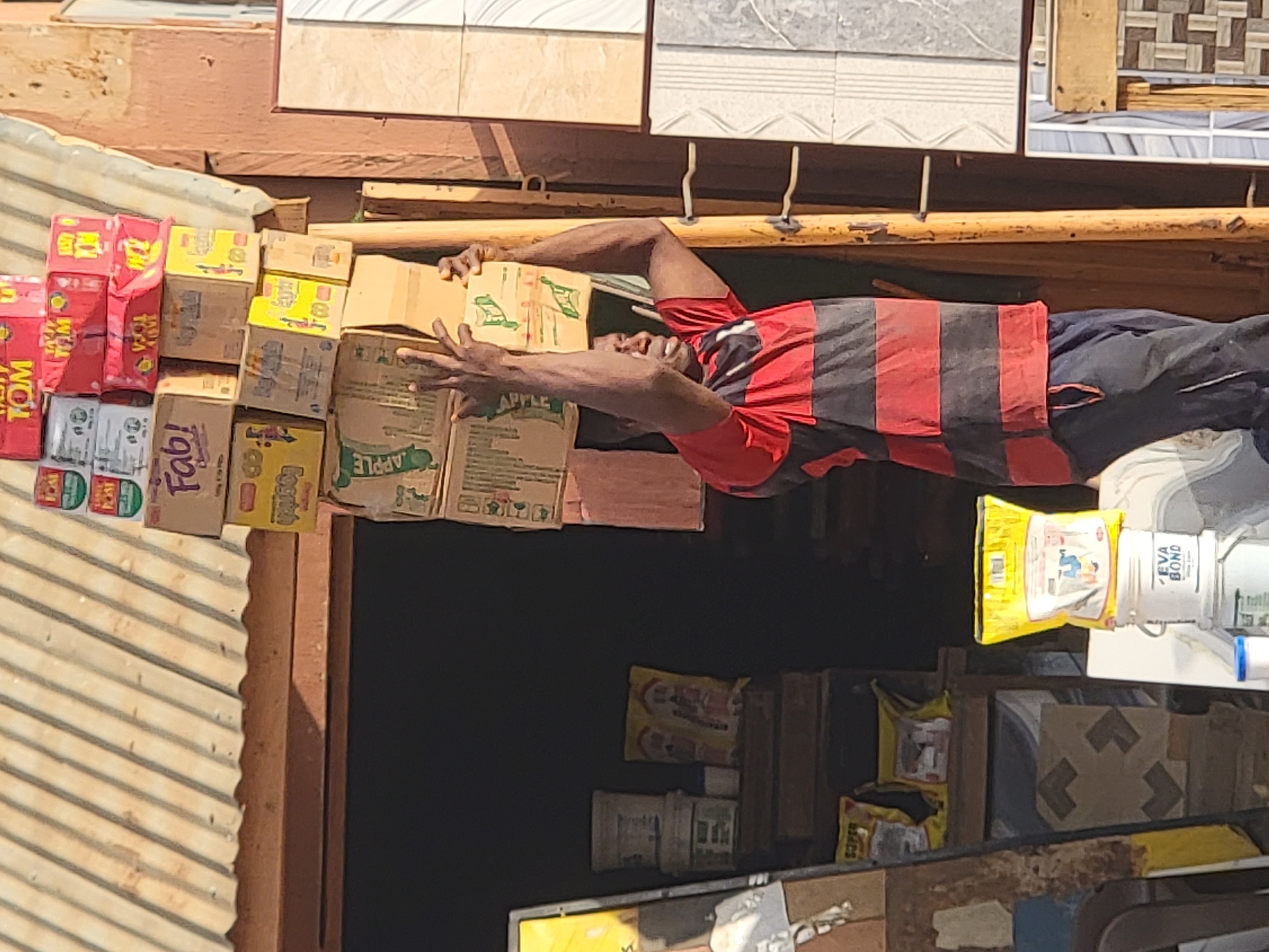 A man in a red and black striped shirt places boxes labeled "APPLE" on a stack of goods in front of a shop in Maiburiji, Nigeria. The shop's inventory includes boxes of "TOM," "Fab!," and other products, as well as construction materials. The storefront is under a corrugated metal awning, and tiles are displayed overhead.