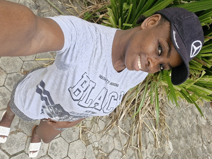 A woman poses for a selfie, wearing a gray t-shirt with "BLACK" printed on it, a black baseball cap adorned with a white Mercedes-Benz logo, and white sandals. She stands on a stone walkway, with green plants and dry grass surrounding her. She smiles slightly, looking directly at the camera. The image is taken from a high angle.
