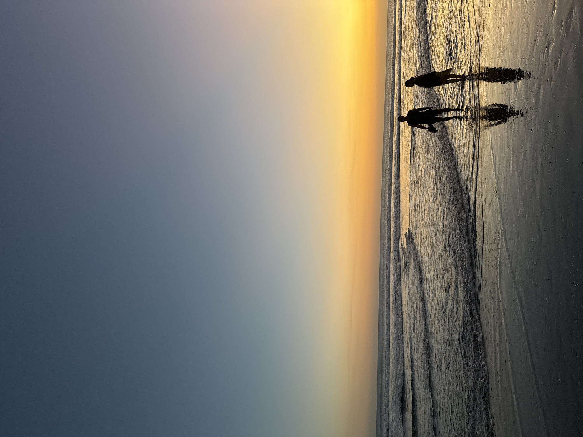 In Kuta, Indonesia, two silhouetted figures, a man and a woman, stand waist-deep in the ocean at sunset. The man, on the left, holds a dark object in his right hand. The woman, on the right, wears a light-colored garment. Their reflections are visible on the wet sand. The sky transitions from a deep blue at the top to a vibrant orange and yellow near the horizon, where the sun has set. Gentle waves lap at the shore, creating ripples on the water's surface and the wet sand. The overall scene is serene and bathed in the warm glow of the setting sun.