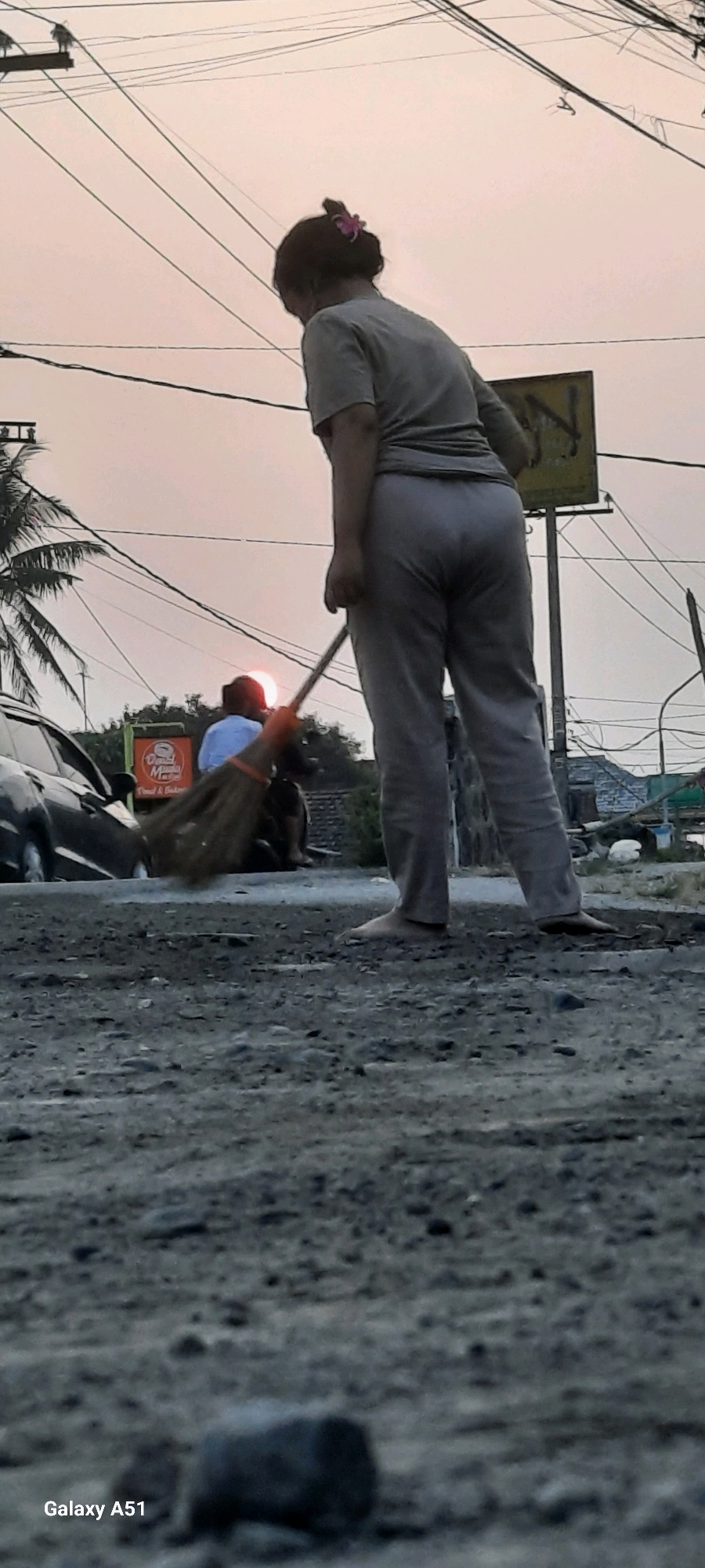 In Selopuro, Indonesia, a person is sweeping a dirt road in the early evening. The sky is overcast with a pale orange hue, suggesting sunset. Overhead, a complex network of electrical wires crisscrosses the sky, supported by poles. The person, viewed from behind, is wearing a grey t-shirt and grey pants, with their hair pulled up and adorned with a pink clip. They are barefoot and holding a broom made of natural fibers, actively sweeping debris on the ground. In the background, a dark-colored car is parked on the left side of the road. Further back, a person on a motorcycle is visible, with the setting sun partially obscuring them, creating a lens flare effect. A sign with red and white text, possibly a business advertisement, is also present in the mid-ground. The ground is uneven and covered with dirt and small stones. The image is captured from a low angle, emphasizing the rough texture of the road. The bottom left corner of the image displays "Galaxy A51".