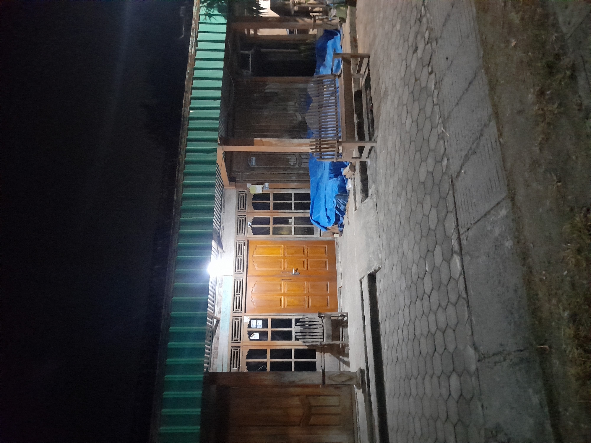The image shows a nighttime scene in front of a building with a green corrugated metal roof. The building has several wooden doors and windows. In the foreground, there is a paved area with hexagonal interlocking concrete pavers. To the left, there is a wooden chair. In the center and to the right, there are blue tarps covering unseen objects. A wooden bench sits on the paved area to the right of the tarps. The overall impression is of a residential or commercial building exterior at night. This location is in Pulokulon, Indonesia.