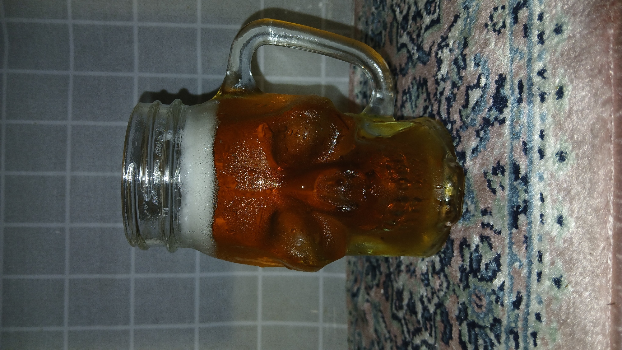 A horizontally oriented, medium shot depicts a skull-shaped beer mug filled with a light amber liquid topped with a white, foamy head. The mug has a clear glass handle. The mug rests on two different surfaces: the left side of the mug sits on a light gray surface with a grid pattern, while the right side rests on a carpet with a busy pattern of blues, grays, and pinks. The scene appears to be indoors, but the location in the city of Unknown, Unknown cannot be determined from the image.