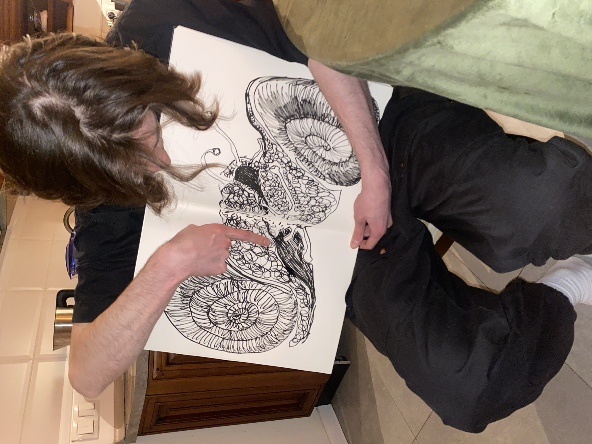 A person with brown hair is holding a drawing of two snails. The person is pointing at the lower snail. The person is wearing black pants, a black shirt, and white socks. The scene appears to be indoors, with kitchen cabinets and a countertop visible in the background. Moscow, Russia is not visible.