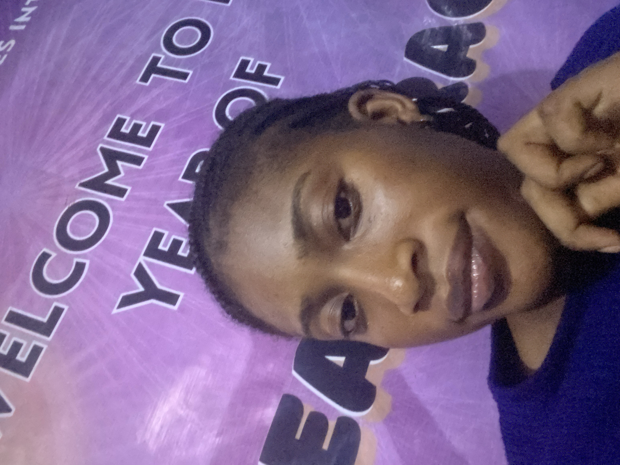 A young adult female, identified as being in Kubwa, Nigeria, is depicted in a close-up, chest-up shot against a purple banner backdrop. She has a dark complexion, dark eyes, and dark, possibly braided hair pulled back from her face. Her lips appear full with a glossy finish. She is wearing a dark blue long-sleeved top and small hoop earrings. Her left hand is positioned near her chin, with fingers slightly curled. The background banner is purple with radiating lines and features large, dark letters outlined in white. Visible text fragments include "WELCOME TO," "YEAR OF," and "GRACE," with additional partially visible letters "ES INT" in the upper right.