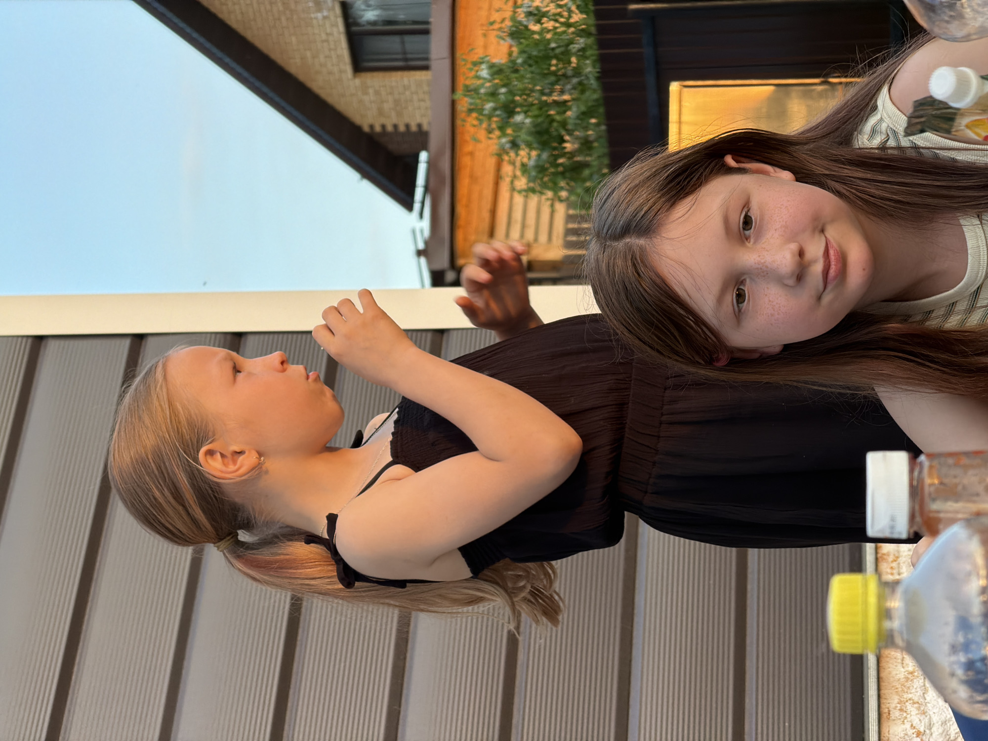 Two girls are positioned in front of a fence. One girl, with blonde hair pulled back in a ponytail, is wearing a black dress. She is looking to the side with her hand near her mouth. The second girl, with long brown hair, is wearing a striped shirt and is looking directly at the camera. Part of a building and a tree are visible in the background. Bottles are positioned on the right side of the image. The location is Chaikovsky, Russia.