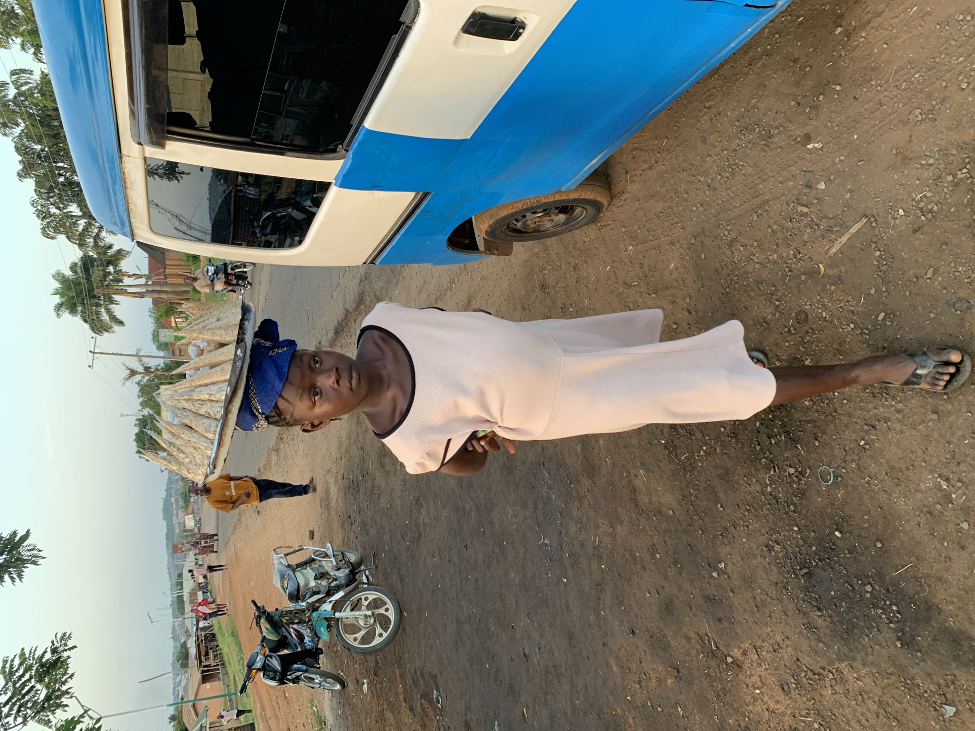 In Gbeleko, Nigeria, a person with a blue headscarf is standing on a dirt road, carrying a load of goods on their head. The person is wearing a light pink dress with a black trim. A blue and white van is partially visible in the upper right of the frame. Two motorcycles are parked on the road. In the background, other people are present, along with buildings and trees under a hazy sky.