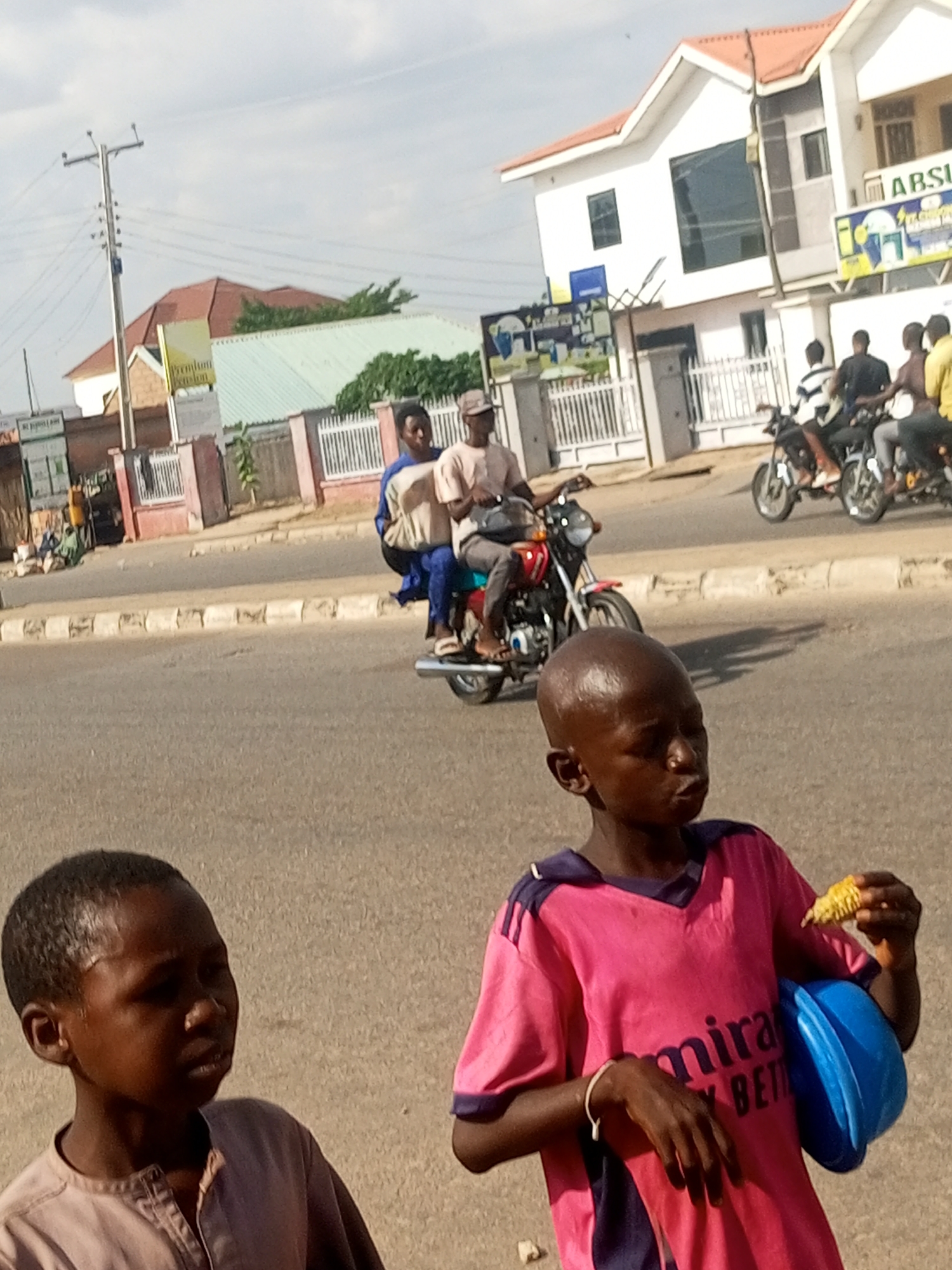 In Pantame, Nigeria, on a sunlit day, a scene unfolds on a paved road. Two young boys are in the foreground, the one on the left wearing a light brown shirt, and the one on the right in a vibrant pink shirt, holding a piece of corn and a blue bowl. Behind them, two motorcycles are prominent. One motorcycle has three people riding on it, with the rider wearing a beige cap and shirt, and two passengers behind him. The second motorcycle, further back, has two riders, one wearing a dark shirt and the other a striped shirt. In the background, there are several buildings, including a white, two-story structure with a red roof and large windows, and a smaller building with a green roof. A traffic light pole stands on the left side of the image. The environment appears to be a busy urban street with signs and fences visible.