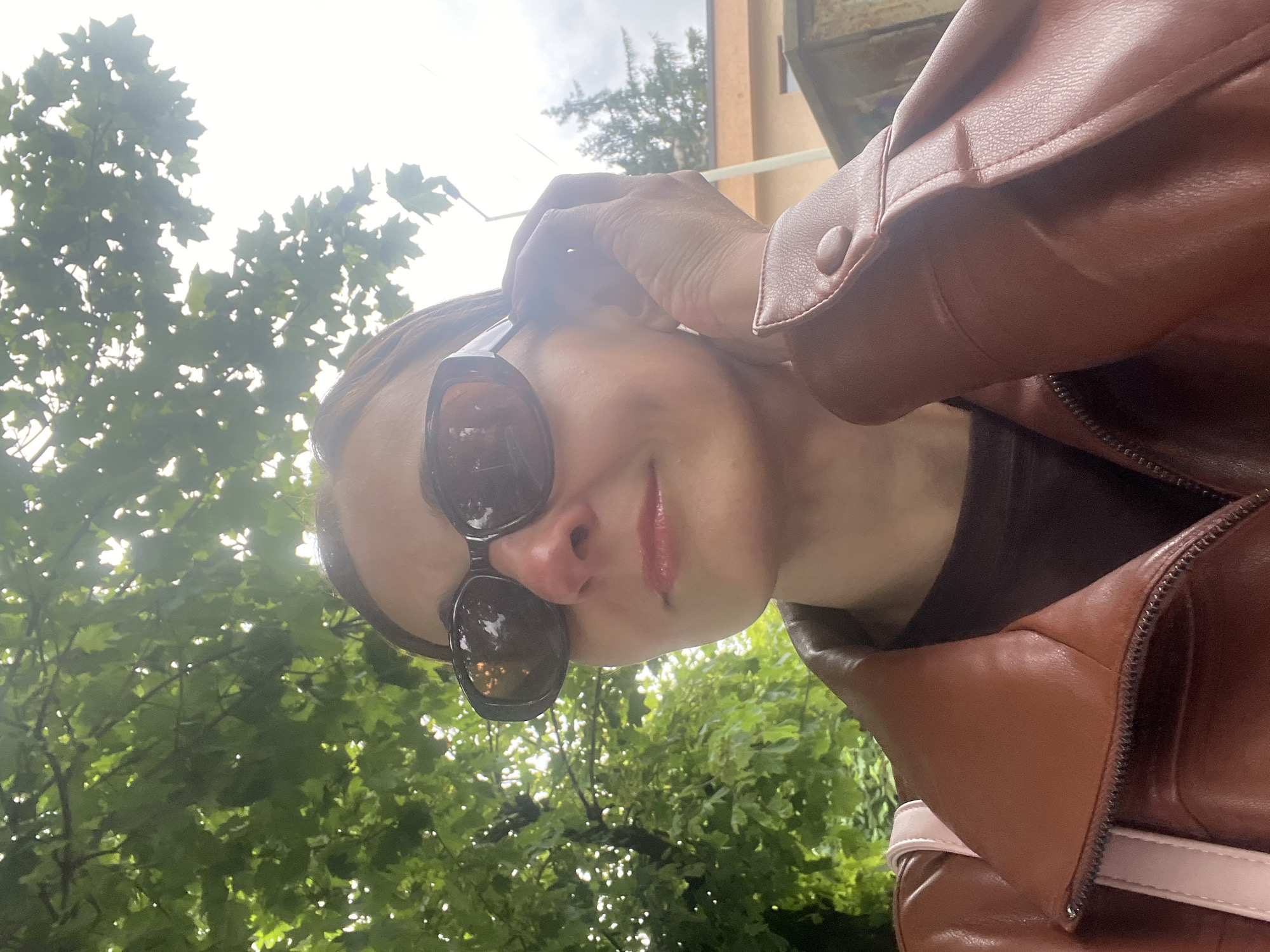 In an outdoor, medium-shot, eye-level, medium-format, vertical, daylight photograph taken in Unknown, Unknown, a woman with fair skin, brown hair, and sunglasses poses for a selfie. She is wearing a brown leather jacket over a dark-colored shirt and a pink strap from a backpack is visible. Her right hand is raised to her ear, and she is looking to her left. Behind her, green foliage and a cloudy sky are visible, along with a building and a lamp post.