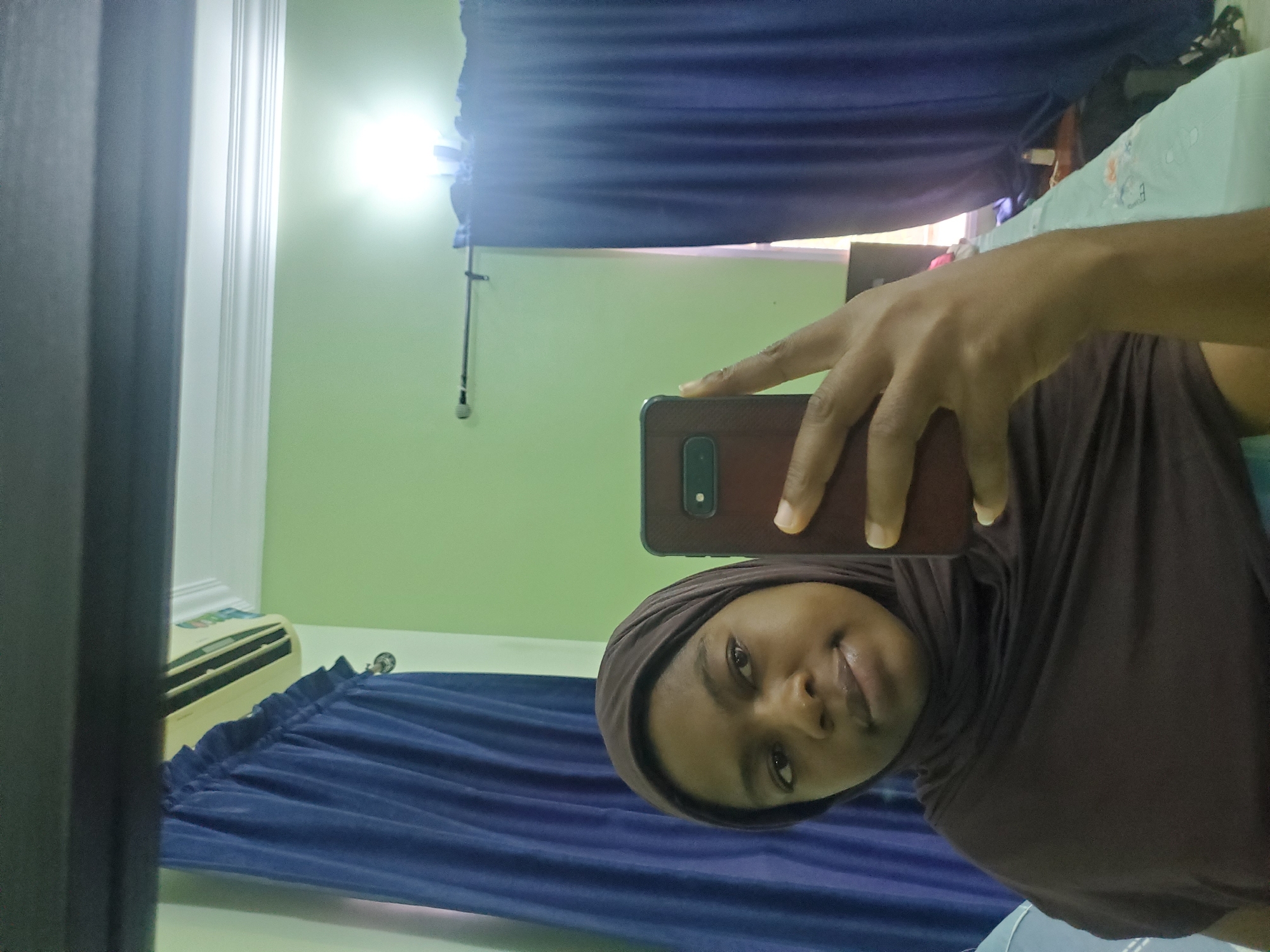 A person wearing a brown head covering holds a smartphone in their right hand, taking a selfie in a room. The room has light green walls and blue curtains. An air conditioning unit is visible in the background. The location is likely an interior space in Barden Yana, Nigeria.