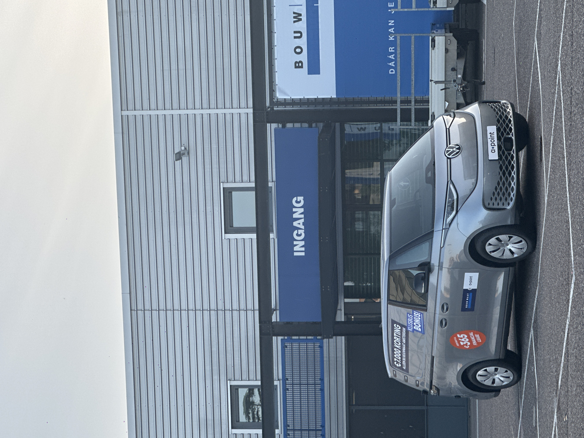 The image shows a silver Volkswagen ID. Buzz parked in a parking lot in front of a modern building. The building has a metallic, corrugated facade and a large blue banner with the word "INGANG" (entrance) in white letters. To the right of the entrance banner is another large banner with blue and white sections and the text "BOUW" and "DÁÁR KAN JE". The van has several stickers on its side, including one that says "€7.000 KORTING" (discount) and another with "$365 FINANCIAL LEASE". The parking lot is paved with white lines indicating parking spaces. A small trailer is visible behind the van. The sky is overcast. This scene is located in Amsterdam, Netherlands, specifically at Bouwmaat Amsterdam, as indicated by the text on the van. There are no people visible in the image.