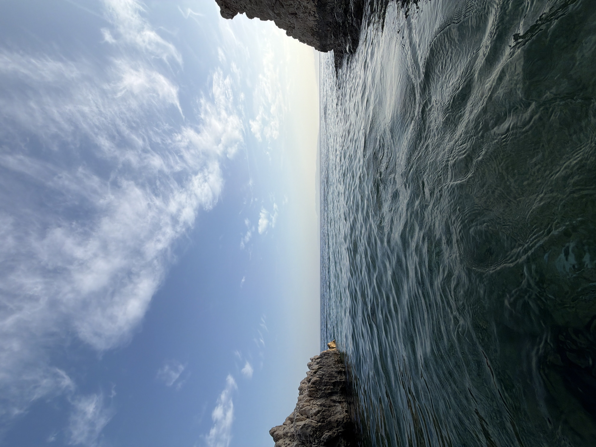 The image displays a serene coastal landscape under a bright daytime sky. The scene is dominated by a clear blue sky and a calm body of water, likely the sea, separated by a distinct horizon line.
The upper portion of the image features an expanse of blue sky, transitioning from a deeper hue at the top to a lighter, almost hazy white-blue closer to the horizon. Scattered across the sky are thin, wispy white clouds, appearing as streaks and patches, indicating fair weather.
The lower half of the image is filled with dark, deep blue-green water, which appears calm with subtle ripples and textured waves reflecting the light. The water extends towards the distant horizon.
Framing the bottom right of the image is a dark, rugged rock formation or cliff edge, showcasing a rough texture. Closer to the center, a smaller, weathered rock outcrop emerges from the water near the horizon. Perched on this smaller rock is a small, indistinct golden or yellowish object, which appears reflective against the darker surroundings.
No human activity or specific event is taking place, presenting a natural and peaceful scene. The time of day appears to be sunny daytime with clear weather. The location is a coastal area, consistent with Grad Rijeka, Croatia. There is no visible text in the image.