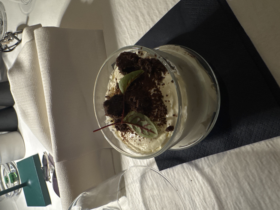 The image displays a dining table setting within a restaurant located in Bolzano, Italy. A clear glass container holds a dessert, consisting of a light-colored creamy base topped with dark chocolate crumbles, three spherical dark chocolate pieces, and two small green leaves with reddish veins, presented on a dark blue paper napkin. To the left, an inverted empty clear glass rests on the white, textured tablecloth. In the background, from left to right, a green stand-like object is partially visible, alongside a clear drinking glass containing liquid and a green item, followed by a white cylindrical container, a folded off-white cloth napkin, two dark cylindrical shakers, and a pair of eyeglasses with dark frames. The scene is static, devoid of people or direct action.
