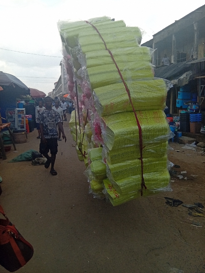 A street-level photograph captures a busy market scene in Ura Nta Umuarandu, Nigeria, under an overcast sky. The prominent subject in the mid-ground is a large, vertically stacked bundle of light-green plastic or paper bowls/containers, each individually wrapped in clear plastic and secured with red ropes. This bundle appears to be in transit, possibly carried by an individual positioned behind it, out of frame. To the left of this bundle, a male pedestrian in a patterned short-sleeved shirt, dark shorts, and sandals walks leftward. Several other indistinct figures are visible further down the street. The ground consists of an unpaved dirt surface with scattered debris. On the left, market stalls with dark canopies display goods including plastic containers and a cooler. On the right, older multi-story buildings with balconies and corrugated metal elements line the street, with plastic buckets and basins displayed outside. A reddish-brown and black bag is partially visible in the lower left foreground.