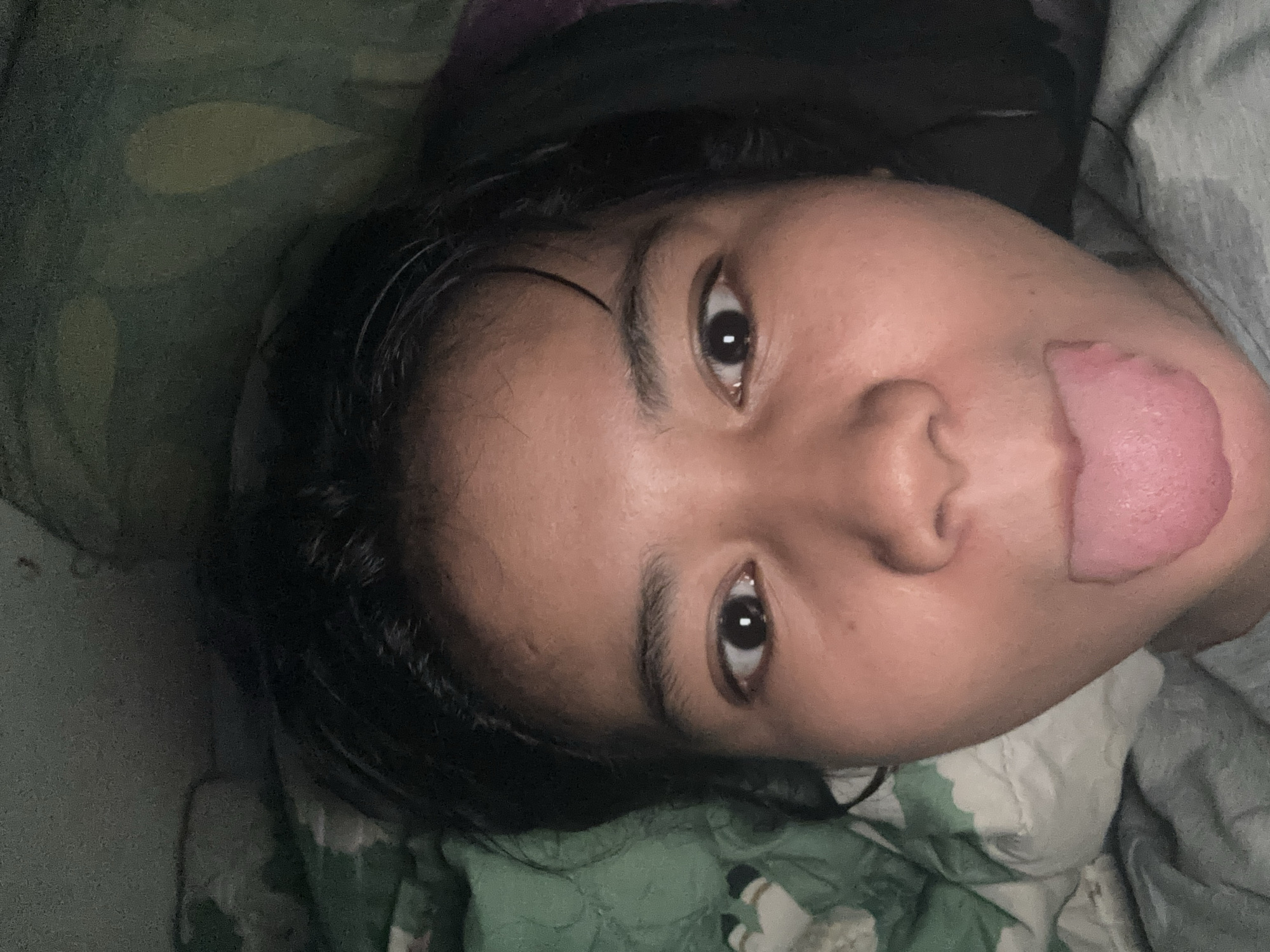 The image shows a close-up, selfie-style shot of a young person's face, with their tongue sticking out. The person has dark hair and is wearing a grey t-shirt. They are lying down, as indicated by the pillows and bedding visible in the background, which appear to be green and white patterned. The lighting is dim, suggesting the photo was taken indoors, possibly at night or in a low-light environment. The context of North Jakarta, Indonesia, does not provide specific details about the scene itself, as it is a general geographical location and does not offer visual cues within the image.