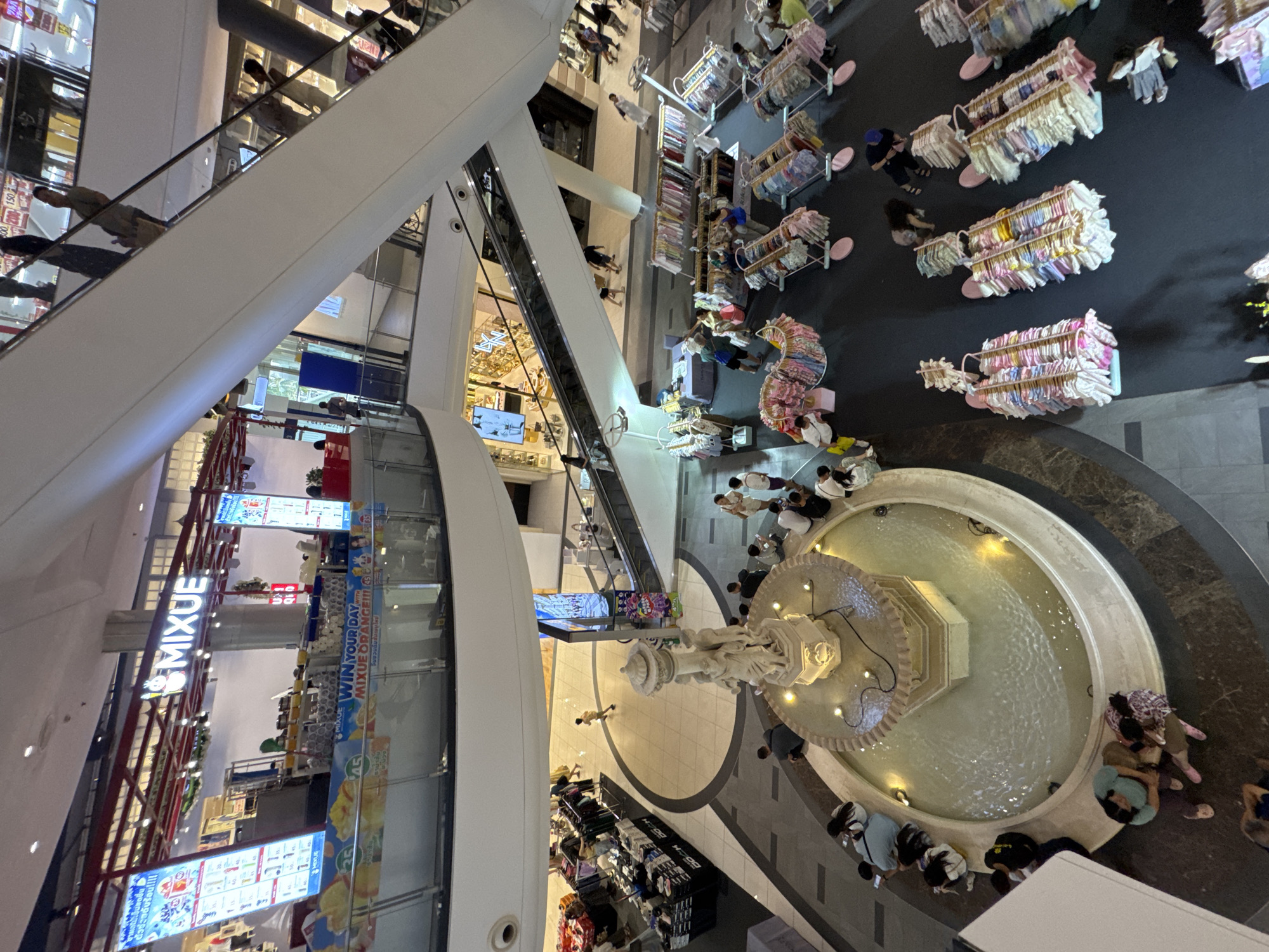 This is a high-angle, full-shot photograph taken inside a multi-level shopping mall in Pattaya City, Thailand. The scene depicts a bustling commercial environment with various activities taking place across different levels.

In the foreground, a large, circular fountain with a decorative sculpture at its center is visible. Several people are seated around the edge of the fountain, some appearing to be resting or conversing. The water in the fountain is illuminated by warm lights.

Moving further back, a wide area of the mall floor is covered with merchandise displays. These appear to be stalls or temporary pop-up shops selling clothing, with racks of garments and various accessories neatly organized. Several individuals are browsing the merchandise, interacting with vendors, or walking through the shopping area.

The mall features prominent escalators and walkways, indicating multiple levels of retail space. In the background, there are signs and storefronts, including a visible "MIXUE" branded stall with large display screens promoting products. The overall environment suggests a vibrant and active shopping destination.

The lighting is a mix of ambient mall illumination and spot lighting on displays, creating a warm and inviting atmosphere. The photograph captures the dynamic energy of a popular urban commercial hub in Pattaya.