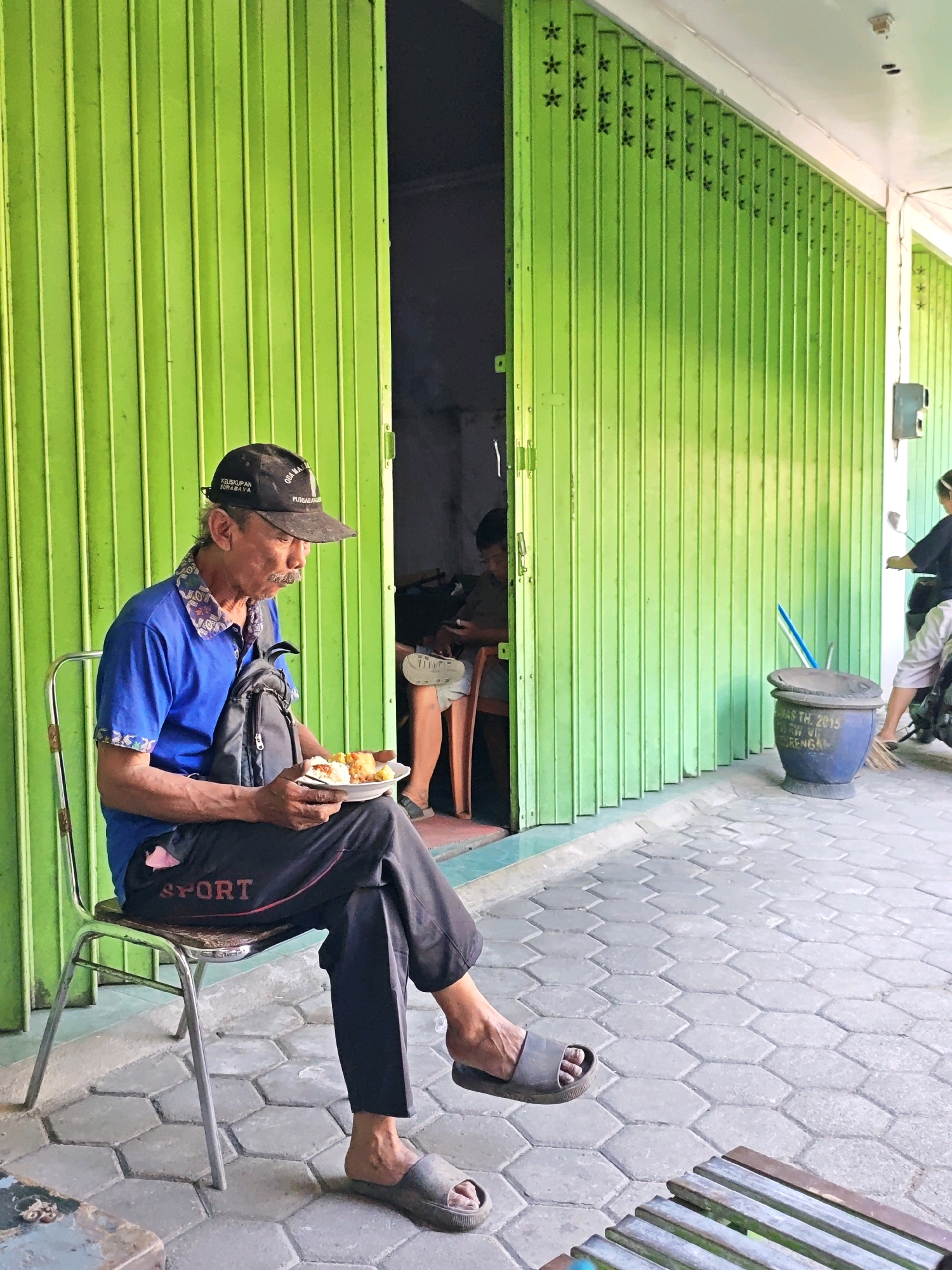 An elderly man with a mustache and a black baseball cap sits on a metal chair, eating from a white plate. He wears a blue polo shirt and black pants with the word "SPORT" in red lettering. The man is positioned in front of a closed, bright green corrugated metal gate. To his right, an open doorway reveals a glimpse of an interior space with another person sitting. To the far right, a blue pot sits on the hexagonal-tiled pavement, and a person on a motorcycle is partially visible. The scene appears to be an outdoor area of a shop or business in Kediri City, Indonesia, given the context of the original prompt.