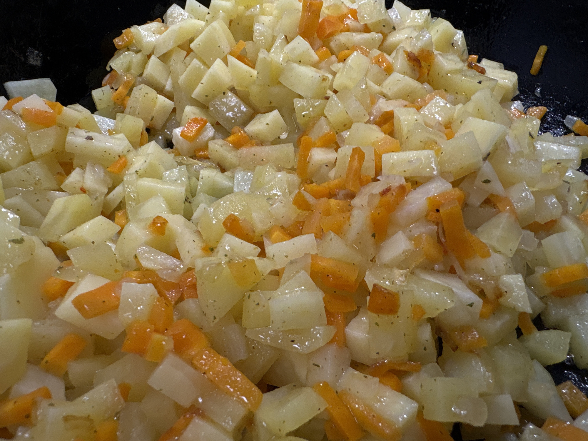 A close-up, overhead shot shows a pile of diced potatoes and carrots in a black pan. The vegetables are glistening and appear to be cooked, possibly sautéed or roasted. Some small green flecks, likely herbs or spices, are visible on the potatoes. The pan is dark, creating a contrast with the lighter colors of the vegetables. The image does not contain any recognizable landmarks or details that would allow for identification of its location within Samara, Russia.