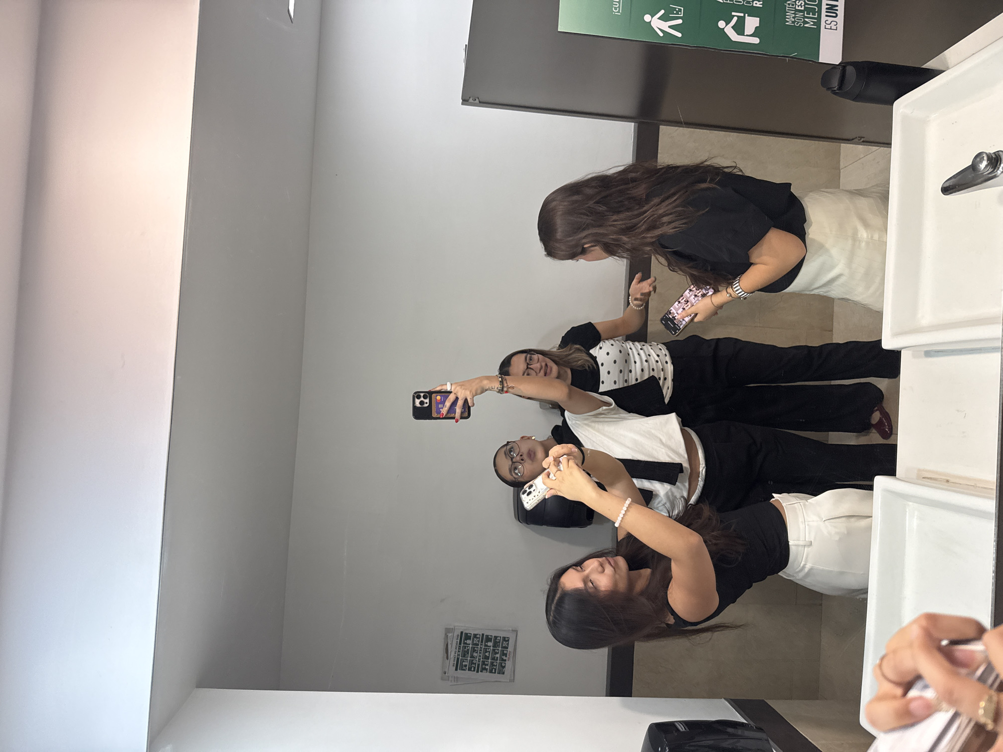 A photo of four women in a bathroom, likely a public restroom, in what can be assumed is San Andrés Cholula, Mexico. The women are taking a selfie in front of a mirror. They are positioned in a line, with the first woman holding up her phone to take the picture. The women are dressed in casual clothing. The restroom has white walls and tile flooring. There are sinks and a mirror visible in the frame. A sign in Spanish is located above the sinks.