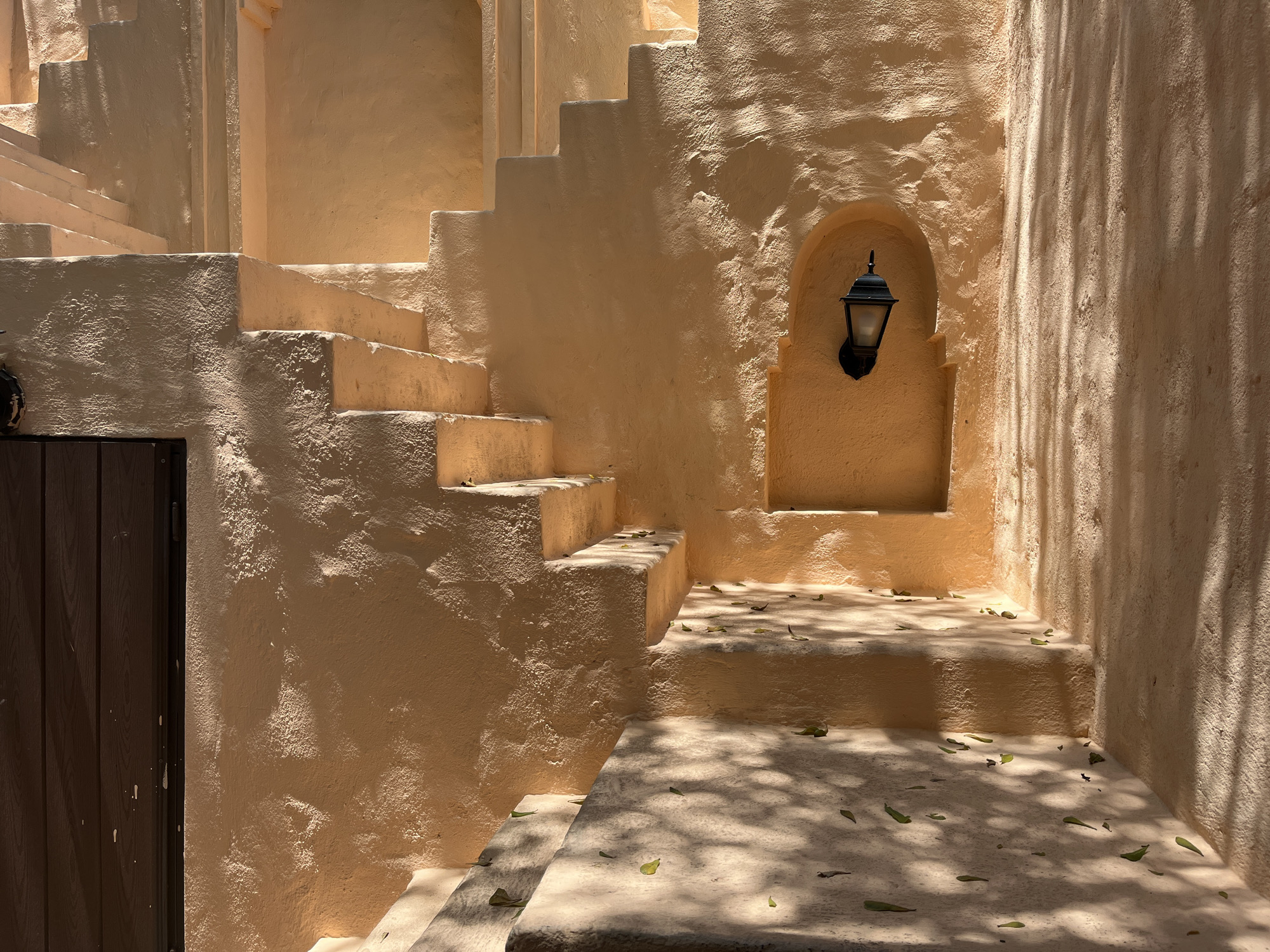 The image shows a sunlit architectural scene with a set of stairs in a building with stucco walls, possibly located in Đà Nẵng, Vietnam, based on the architectural style reminiscent of Mediterranean or Middle Eastern designs sometimes found in Vietnamese tourist areas. A dark wooden door is partially visible on the left. An arched alcove on the right wall houses a black wall-mounted lantern. Sunlight filters through the trees, casting shadows on the walls and stairs, creating dappled patterns on the steps. A few fallen leaves are scattered on the steps. There are no people visible in the scene.