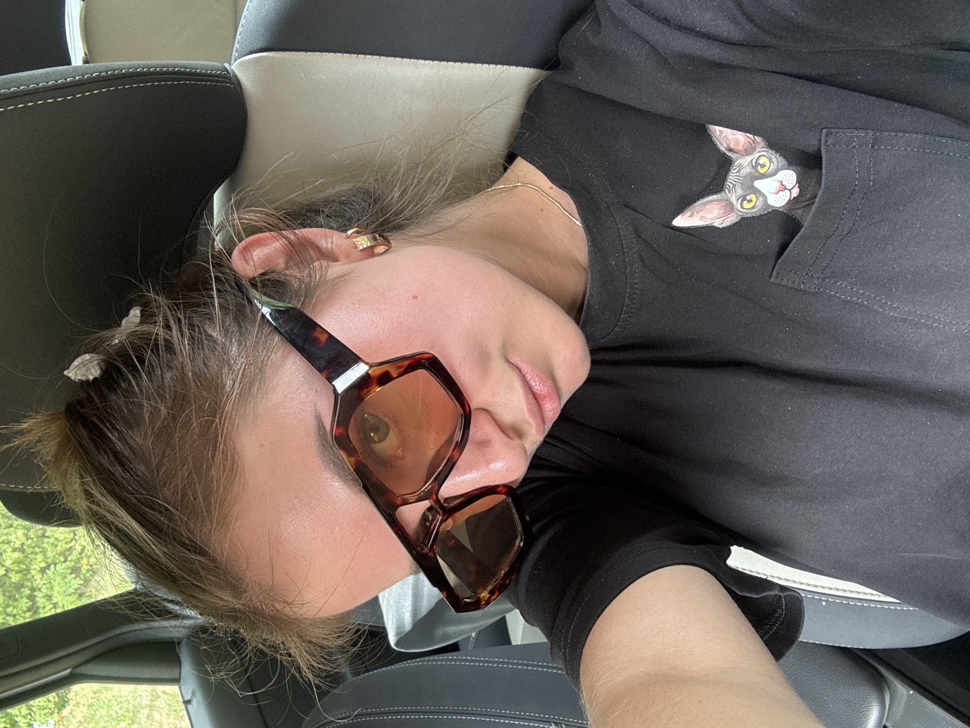 A young woman with her hair pulled up in a bun and wearing large, tortoiseshell sunglasses is in the driver's seat of a car. She is wearing a black t-shirt with a printed image of a Sphynx cat in the breast pocket. The car interior is visible, including a black and grey seat with white stitching. Outside the car, green foliage is visible. The scene appears to be on a bright, sunny day.