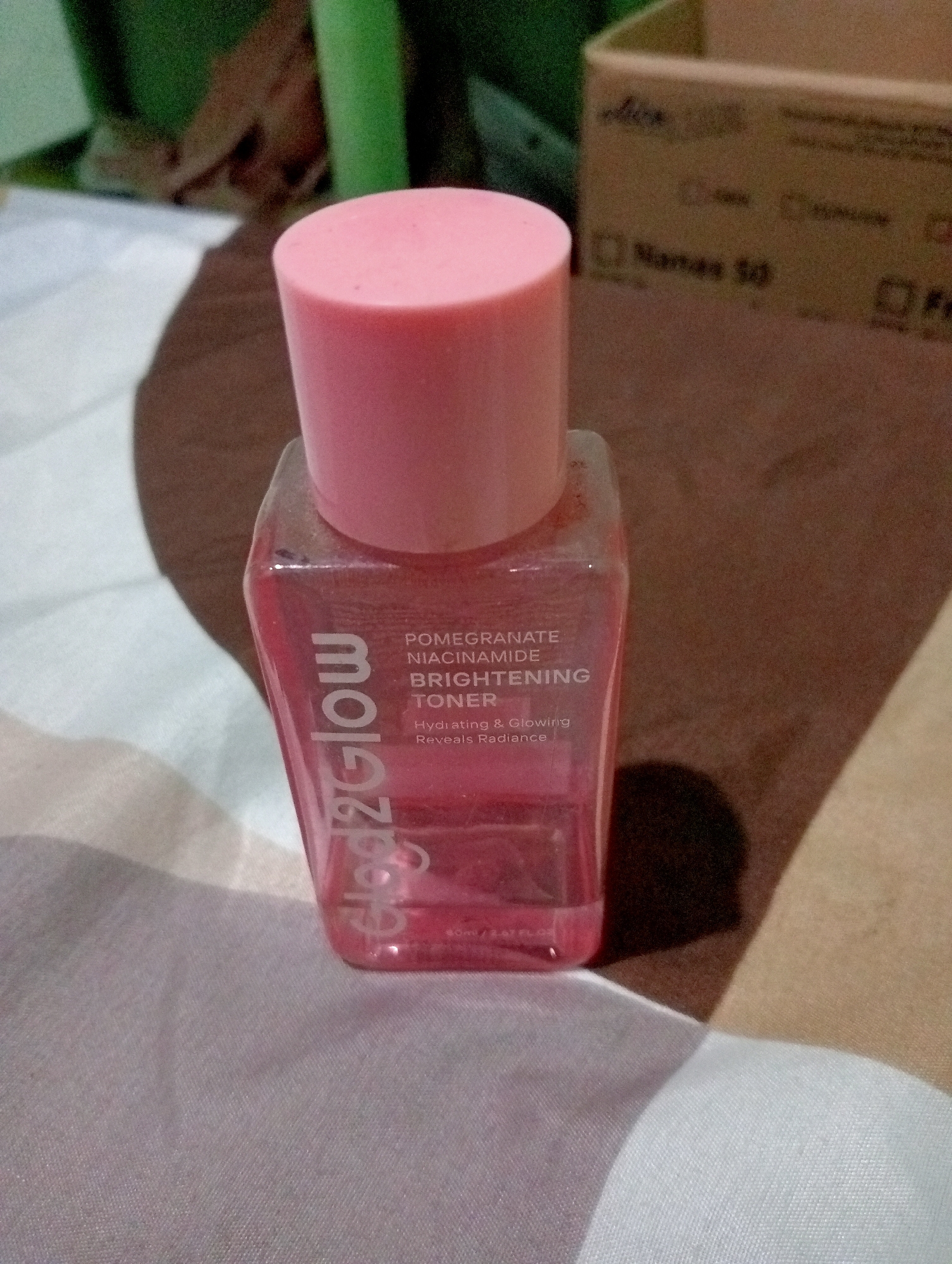 A pink-capped, clear glass bottle of toner with pink liquid is shown in the foreground, with its label facing the viewer. The label reads "Glow2Glow" vertically, and then "POMEGRANATE NIACINAMIDE BRIGHTENING TONER" horizontally. Below that, it says "Hydrating & Glowing Reveals Radiance." The bottle is resting on a brown, textured surface, possibly a bedspread or fabric. In the blurred background, there is a cardboard box with text that is mostly illegible, and some green objects that appear to be plants. The lighting suggests an indoor setting, possibly a bedroom. The image is not taken in the city of Kota Negara Timur, Indonesia; it's a generic indoor product photograph.