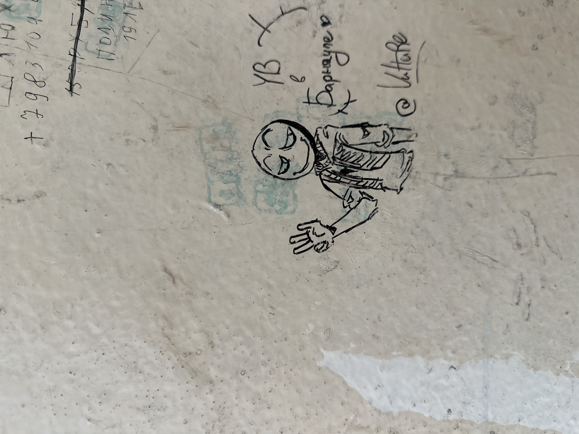 The image displays a section of a textured, light-colored wall, possibly concrete or plaster, marked with various graffiti. In the center-left, a cartoonish drawing of a figure with a wide smile and closed eyes is depicted. The figure wears a hooded jacket and holds its right hand up in a peace sign. To the right of the drawing, graffiti in Russian script is visible, including "УВ в Барнауле" which translates to "UV in Barnaul," and "@ Vatture." Above these writings, other inscriptions are present, some of which are numbers like "+7983104" and "19LE." The overall environment appears to be an outdoor urban setting, likely a public wall in the city of Barnaul, Russia, covered in various forms of street art and inscriptions.