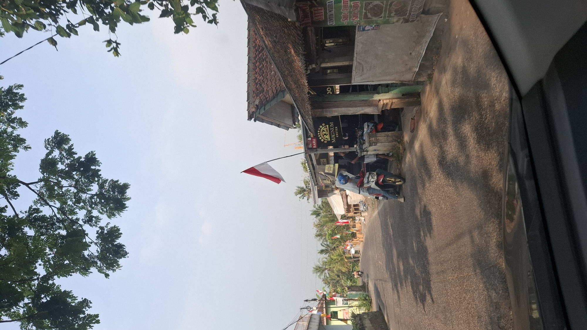 In Cibinong Hilir, Indonesia, a residential street is captured under a bright, hazy sky. Lush green foliage from trees overhangs the scene, casting dappled shadows on the asphalt road. The street is lined with modest, single-story homes with terracotta tile roofs, some displaying small Indonesian flags. In the mid-ground, a red and white Indonesian flag is prominently displayed on a pole in front of a small shop. Two people are visible near a motorcycle parked by the shop, with another motorcycle in the background. The shop appears to be a local vendor, with signage for "MAGNUM KRETEK" and what seems to be a menu board with food items like "Sate Ayam" and "Sate Sapi." The foreground shows the dashboard and hood of a vehicle, indicating the photo was taken from inside a car. The overall impression is a quiet, sunny day in a rural Indonesian community.