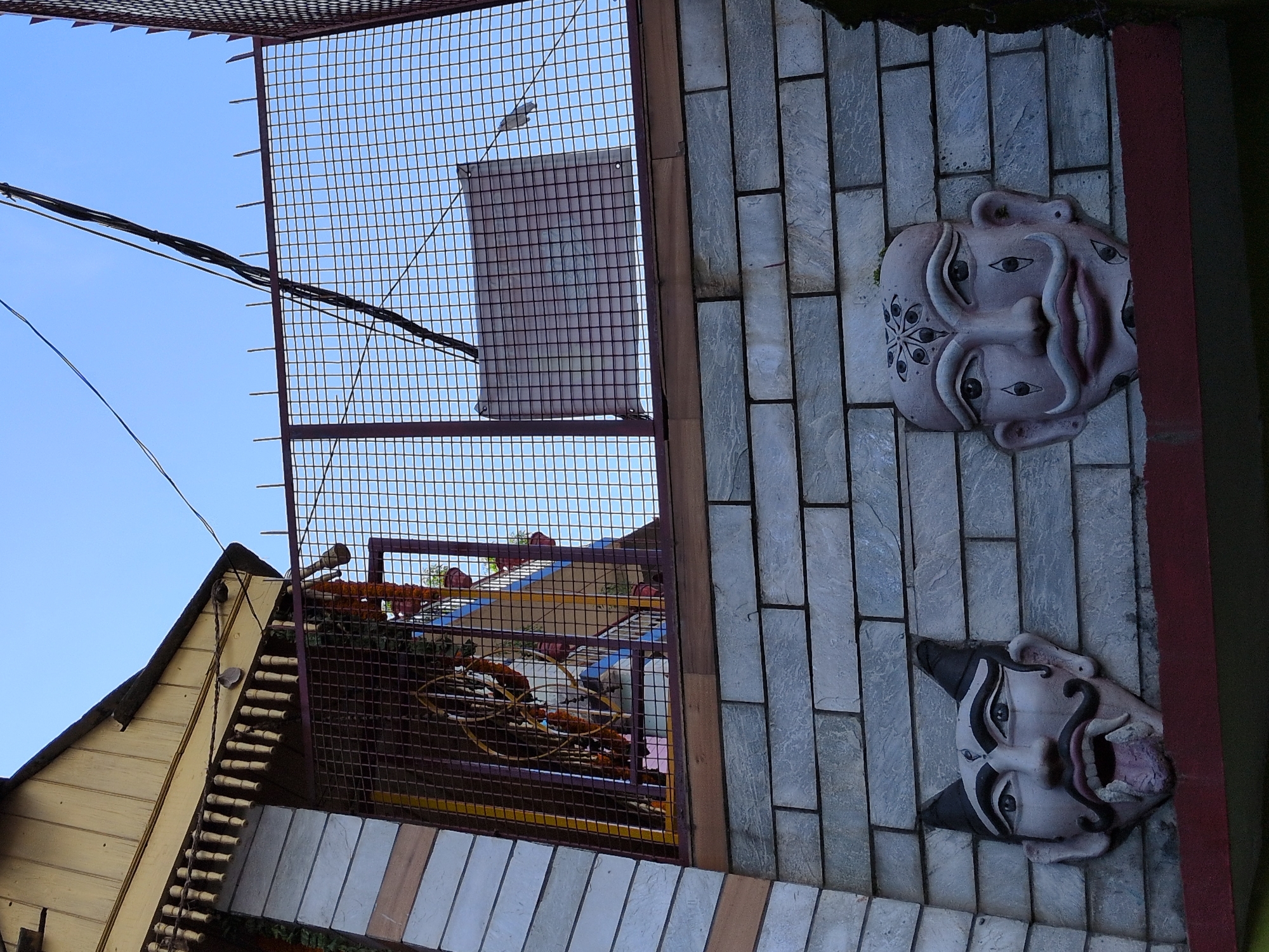 The image shows a street view in Kullu, India, with a building facade featuring decorative masks and a wire fence. On the left, a building section has a light-colored, possibly wooden, overhang with decorative vertical elements. Below this, the wall is constructed with grey, rectangular stone blocks. Two distinct masks are mounted on this stone wall. The mask on the left is a dark-colored, demonic-looking face with pointed ears, a mustache, sharp teeth, and a protruding tongue. The mask on the right is a lighter-colored, human-like face with a mustache and decorative markings on the forehead and cheeks resembling eyes. A wire fence with a reddish-brown frame is visible behind the masks, enclosing an area where some greenery and what appears to be a colorful structure can be seen. Power lines crisscross the sky above the fence. The sky is clear and blue. The overall scene suggests an outdoor setting with elements of cultural or religious decoration.