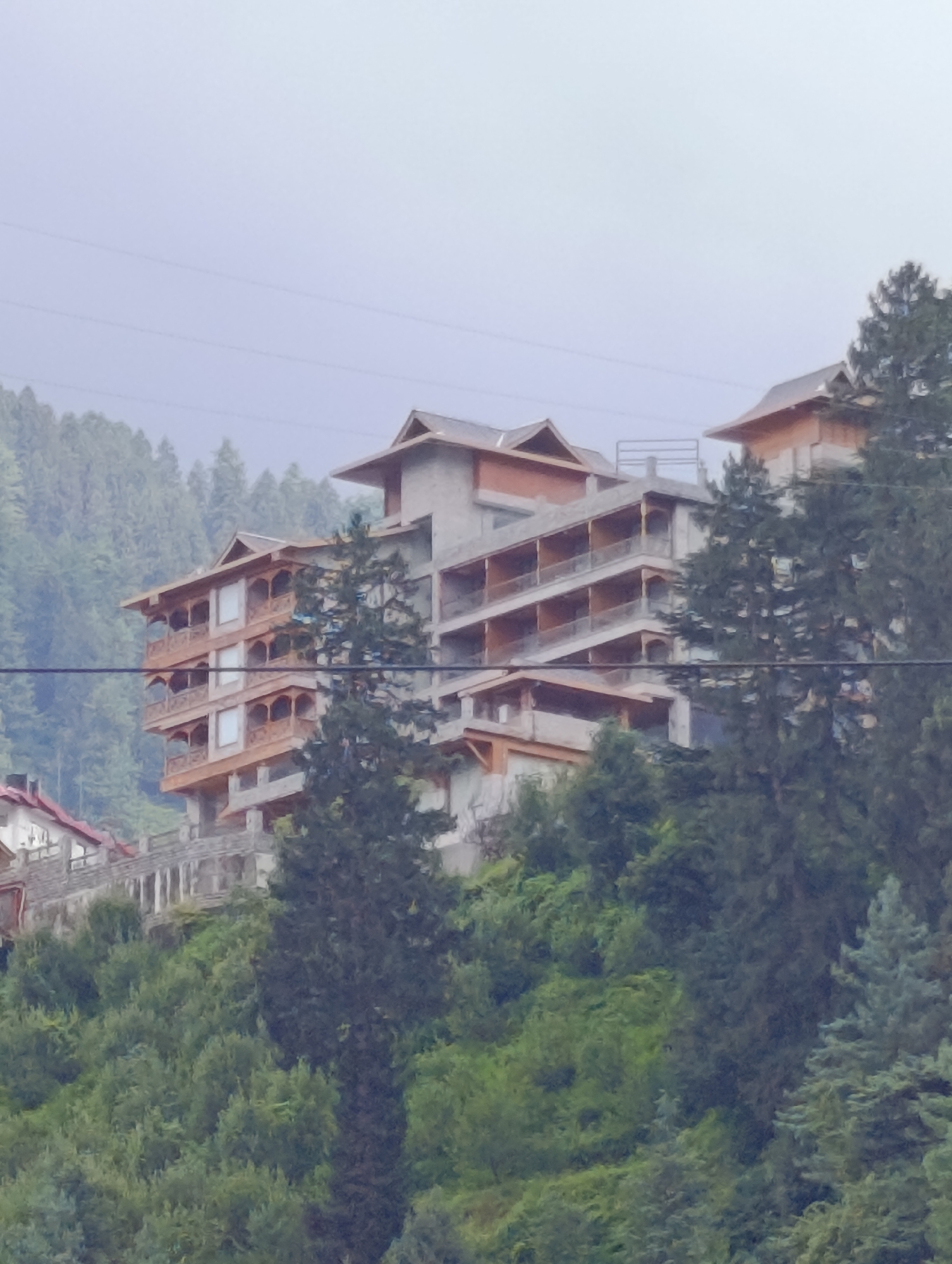 A multi-story building with a unique architectural style, featuring wooden balconies and sloping roofs, is nestled into a lush green hillside. The building appears to be under construction or recently completed, with exposed concrete elements and scaffolding visible. The surrounding environment is dominated by dense coniferous forests, characteristic of a mountainous region. The sky is overcast and hazy, suggesting a cool or misty climate. The overall scene conveys a sense of development in a natural, remote setting, possibly a resort or hotel in a hilly area like Chhyal, India. There are no people or specific actions depicted.