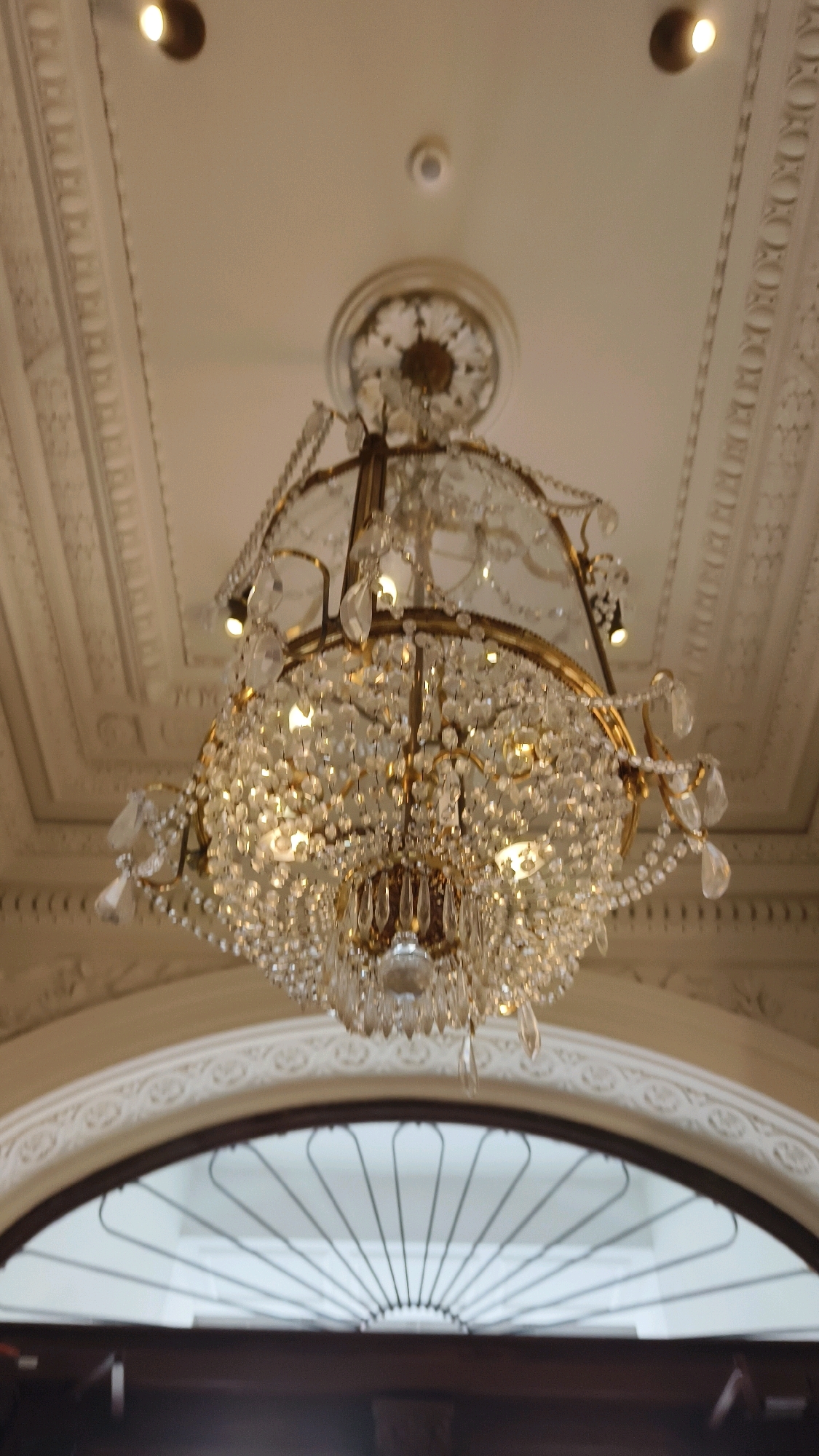 An ornate crystal chandelier hangs from a highly decorated ceiling. The chandelier is made of brass and crystal, with many hanging crystal prisms. The ceiling is white and features intricate moldings, with two recessed spotlights visible in the upper portion of the image. The chandelier hangs above an arched doorway with a decorative metal grate. The scene appears to be indoors, possibly in a grand building such as a museum or historical landmark in Vienna, Austria. No people are visible in the image.