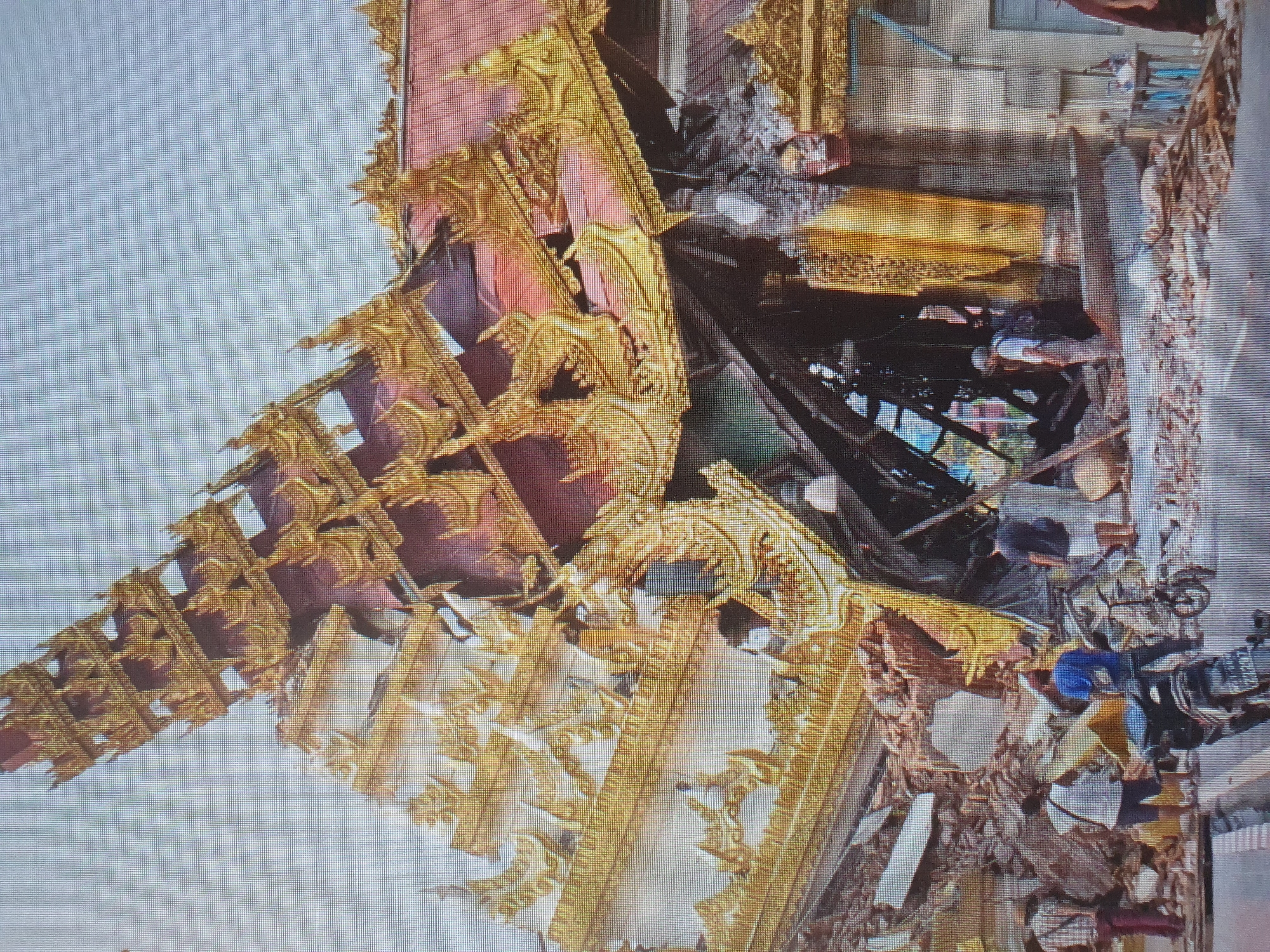 The image depicts the aftermath of a structural collapse, likely a temple or pagoda, given the ornate golden roofs and tiered architectural style. The central structure has severely caved in, with exposed wooden beams and debris scattered around. Several individuals are present, observing the damage. Some are standing near the rubble, while others are in the background. A motorcycle is parked in the foreground. The environment appears to be an urban setting, with a road visible in front of the damaged building. The sky is overcast. The damage suggests a catastrophic event such as an earthquake or structural failure.