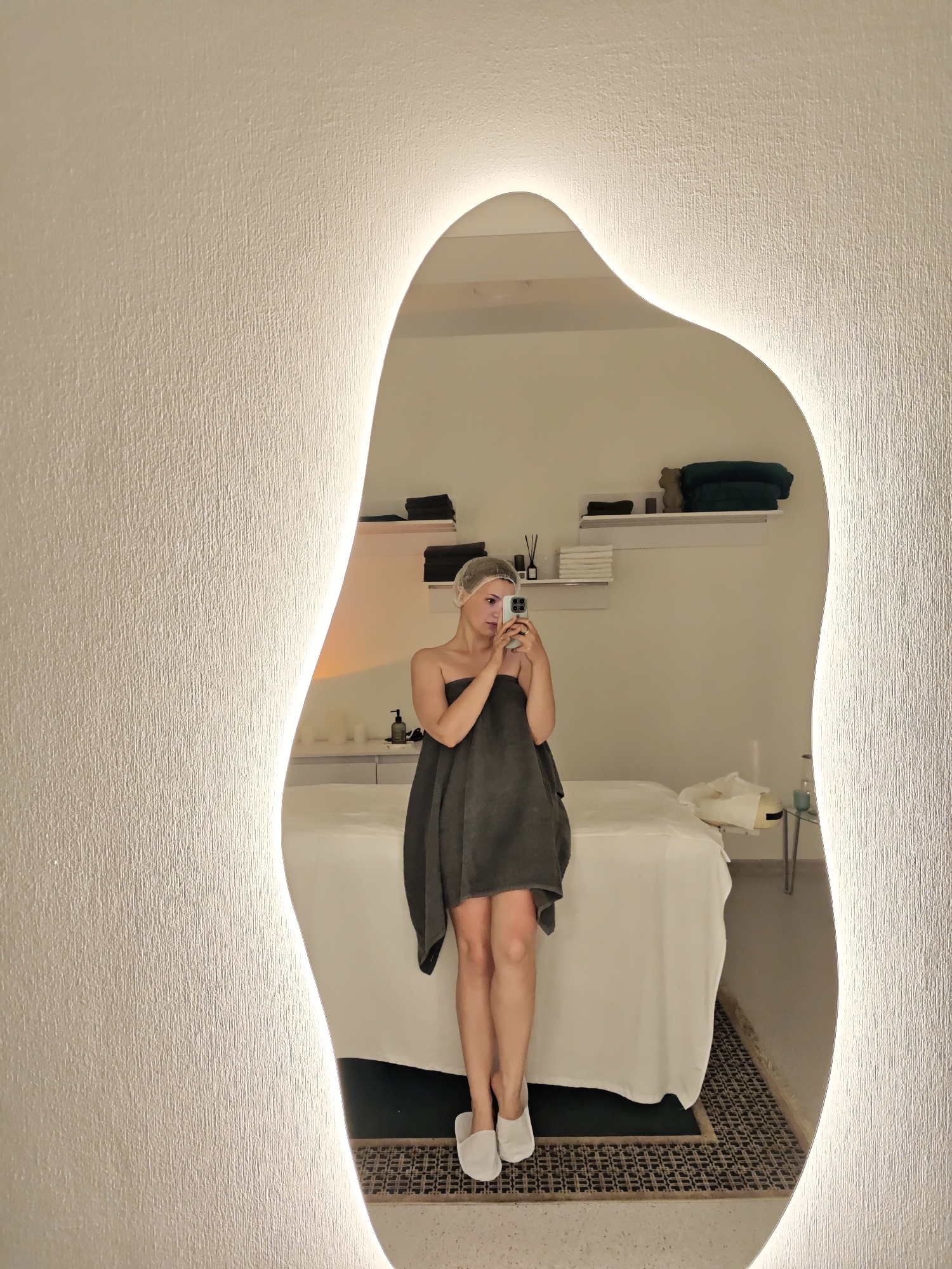 A woman is taking a selfie in a spa located in Vinnytsia, Ukraine. She is standing in front of a mirror with integrated lighting. She is wearing a dark grey towel wrapped around her body and a clear plastic shower cap on her head. She is holding a smartphone in her right hand, capturing the reflection. In the background, a massage table covered with white linens is visible, along with shelves displaying towels and bottles. The room appears to be a treatment room within a spa.