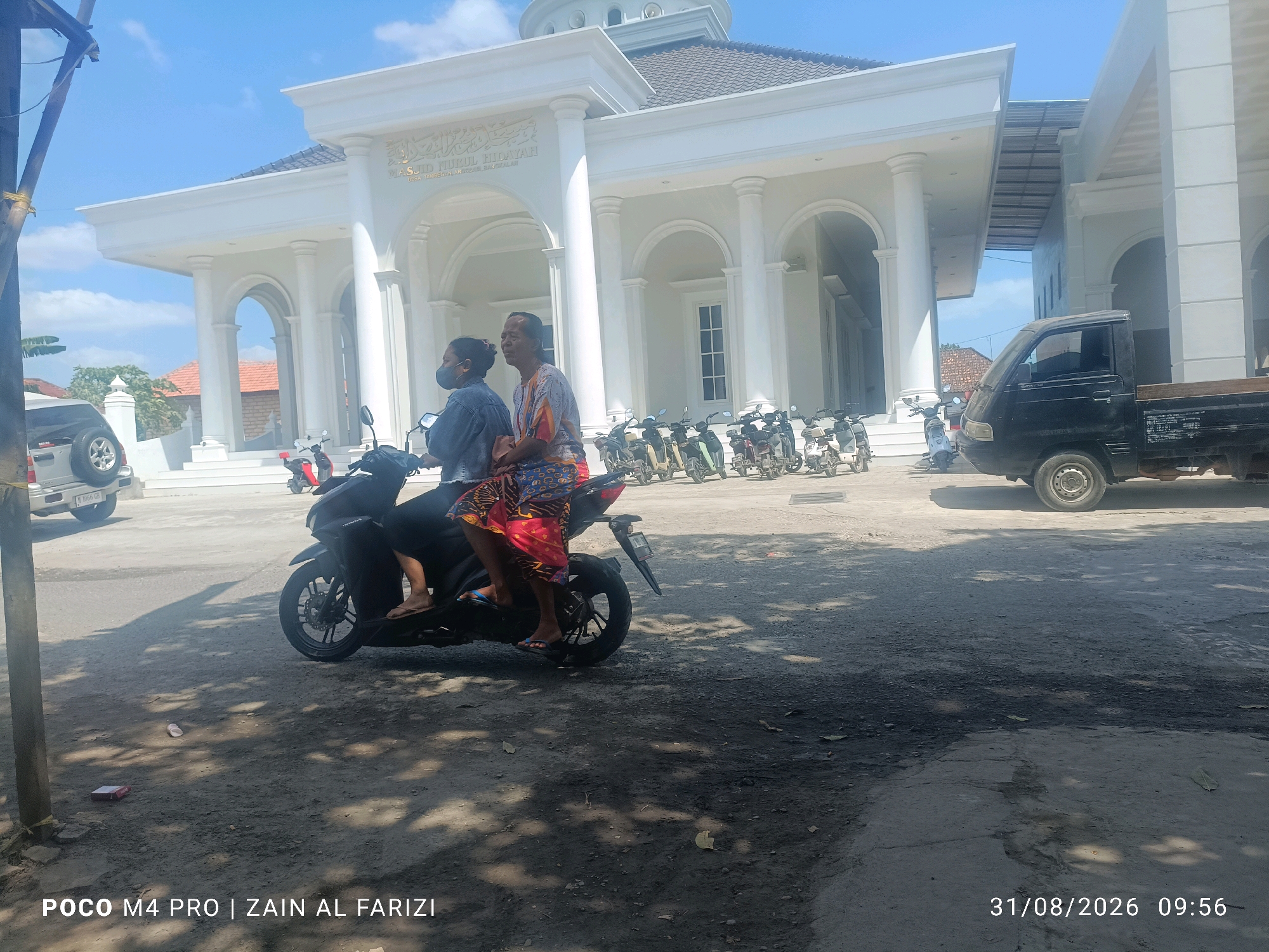 In Cendagah, Indonesia, on a bright, sunny day, a mosque with a white facade and arched entrances stands prominently. Two people, a woman wearing a face mask and a man with a colorful shirt, are riding a black motorcycle. The woman is in the front, and the man is behind her. Parked in front of the mosque are several motorcycles and a black pickup truck. The ground is paved with asphalt and dirt, with shadows cast by trees. A water bottle and a pack of cigarettes are visible on the ground. The image also contains text overlays, "POCO M4 PRO | ZAIN AL FARIZI" and "31/08/2026 09:56".